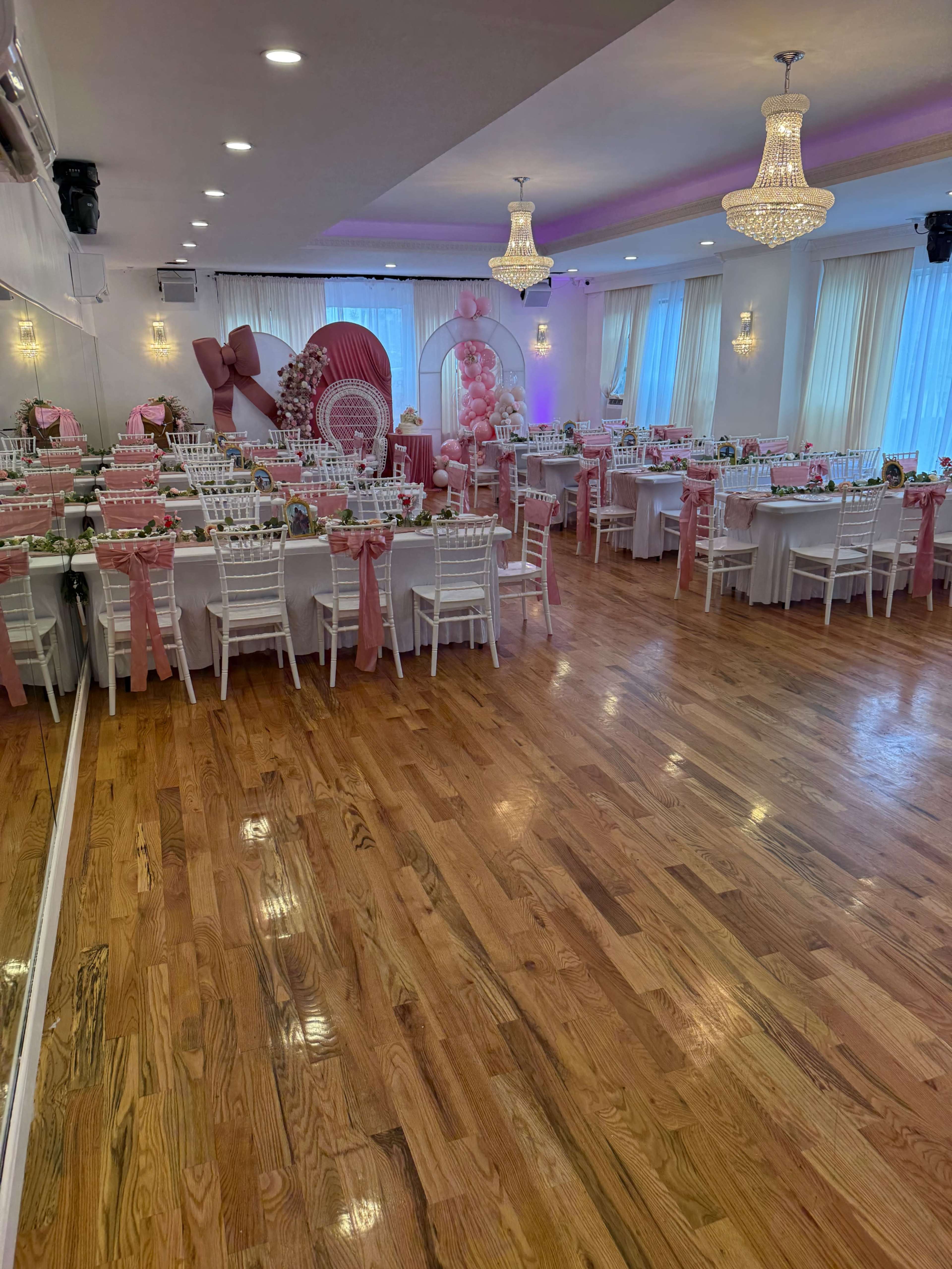 The image shows an elegantly decorated banquet hall with white tables set with pink tablecloths and chairs adorned with pink sashes, surrounded by decorative elements like balloons and chandeliers.
