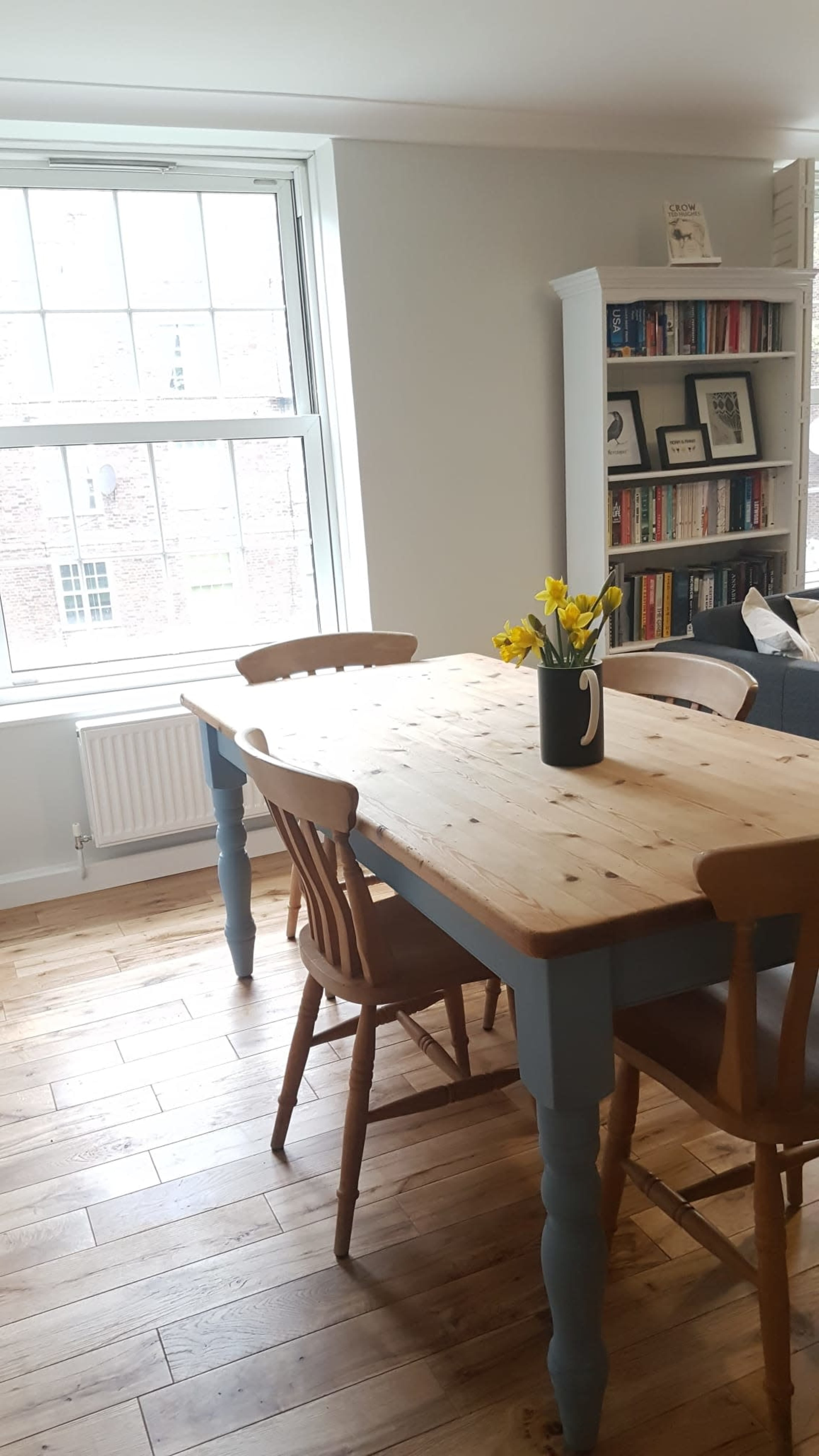 A wooden dining table with light blue legs and two chairs stands in a well-lit room featuring a window and a bookshelf filled with books.