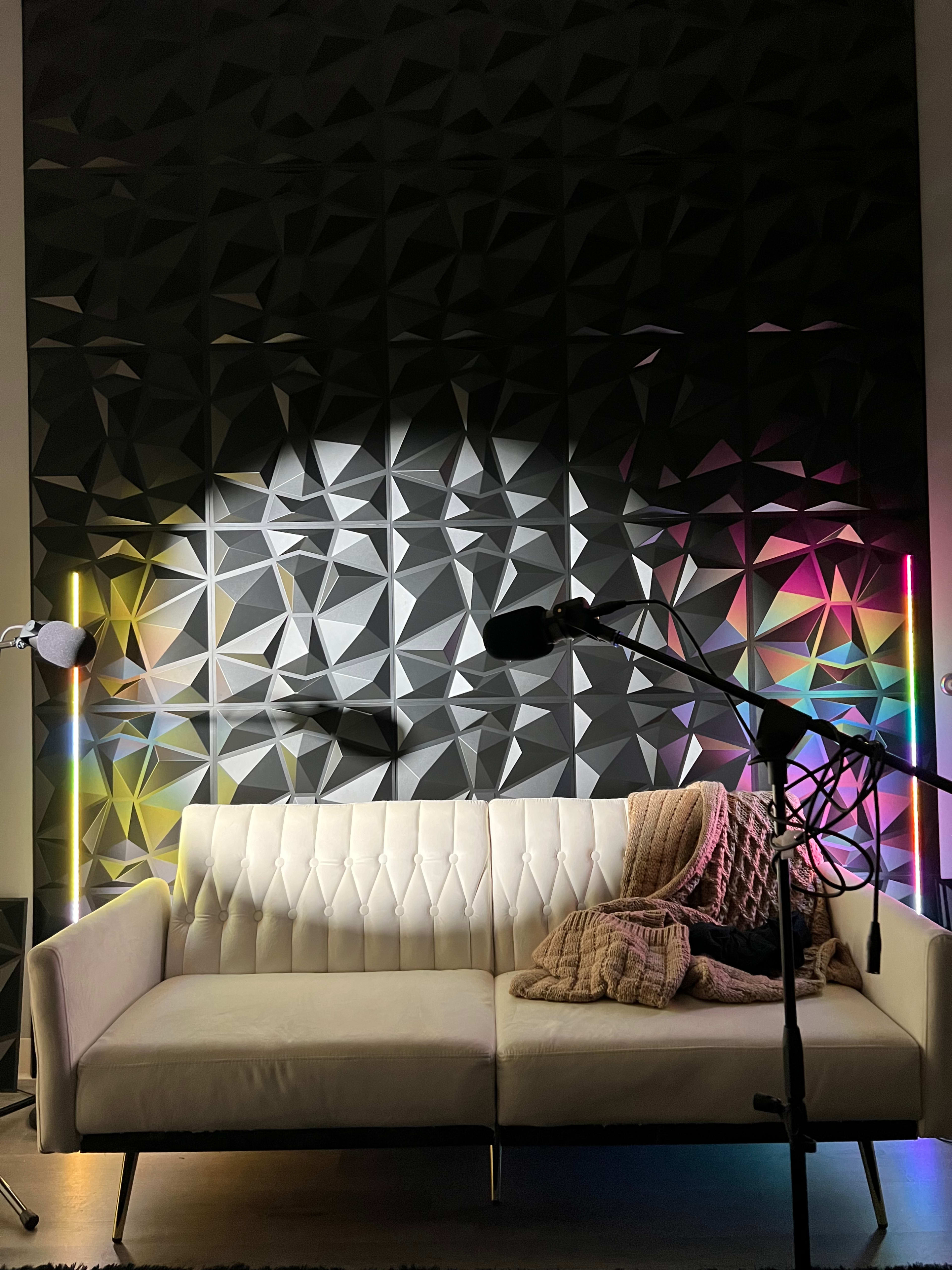 A white sectional sofa is positioned in front of a textured black wall, illuminated by colorful lighting from two standing lamps on either side and a microphone setup nearby.