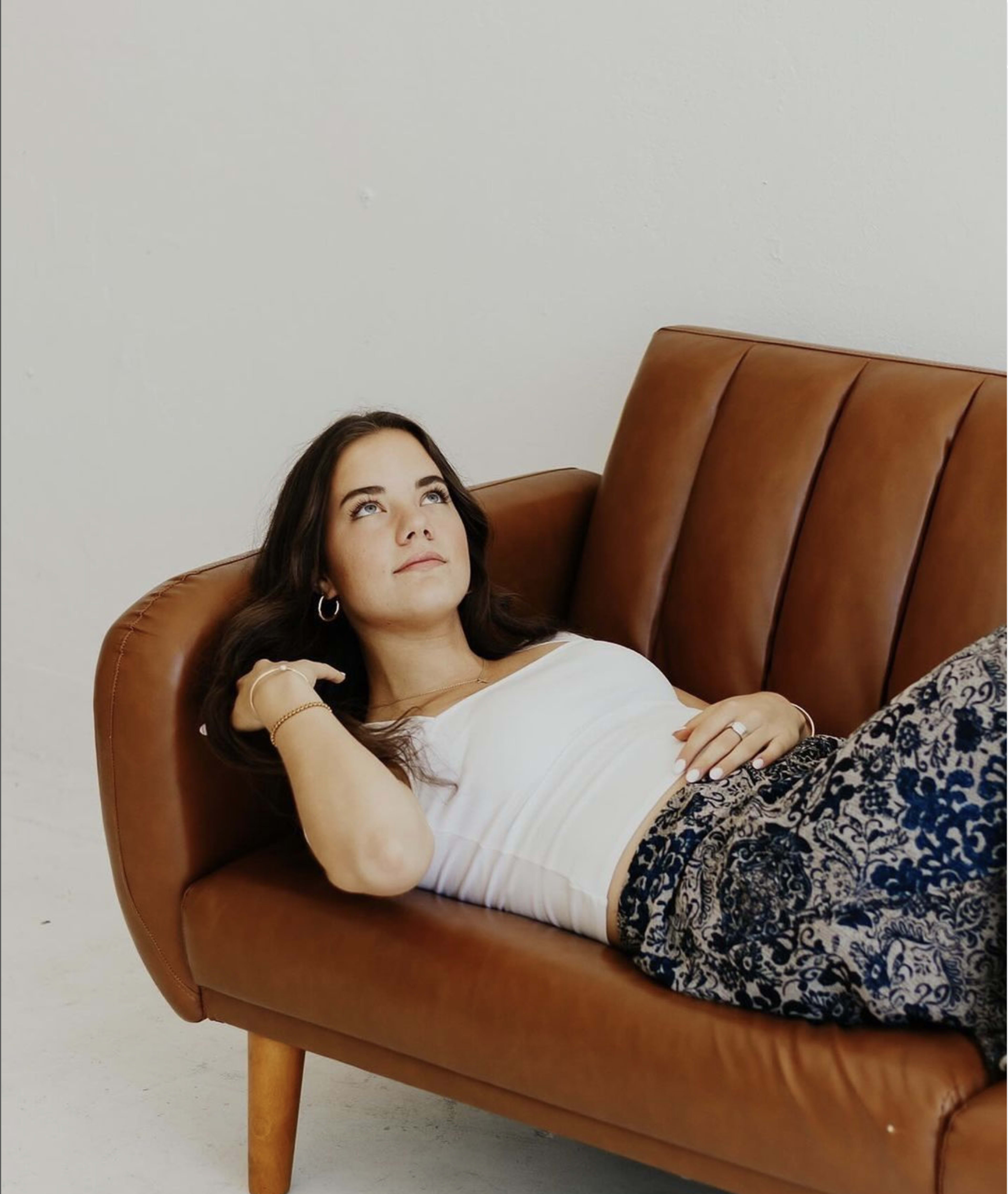 A woman lies on a brown leather couch, looking up thoughtfully with her hand resting behind her head.