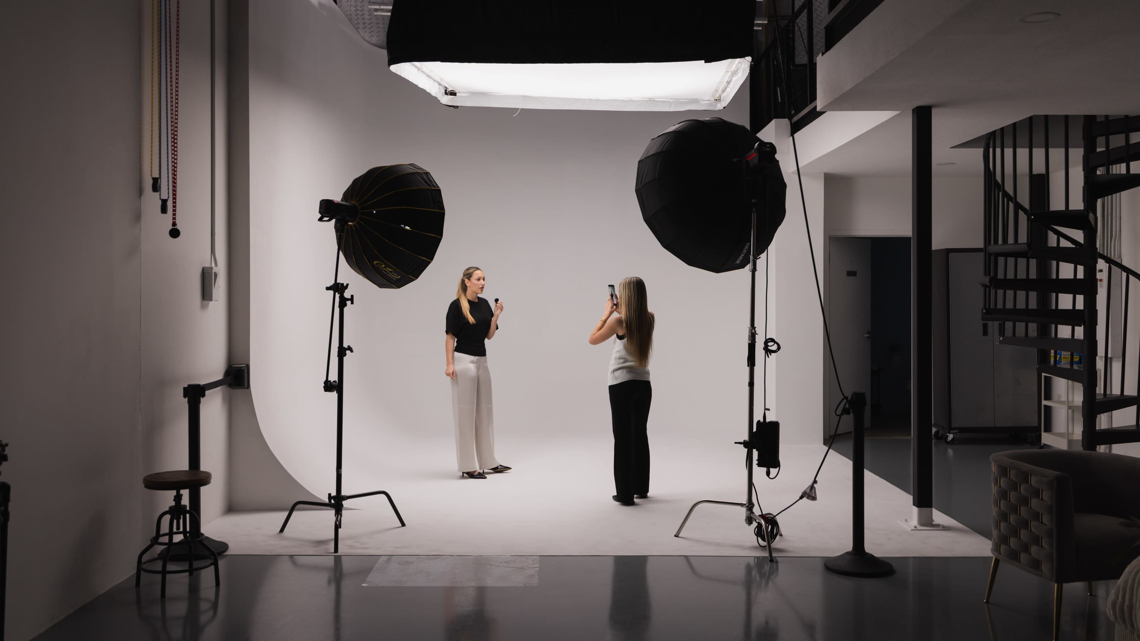 A photoshoot setup features two women, one posing for the camera while the other takes pictures, surrounded by lighting equipment in a studio with a plain white backdrop.