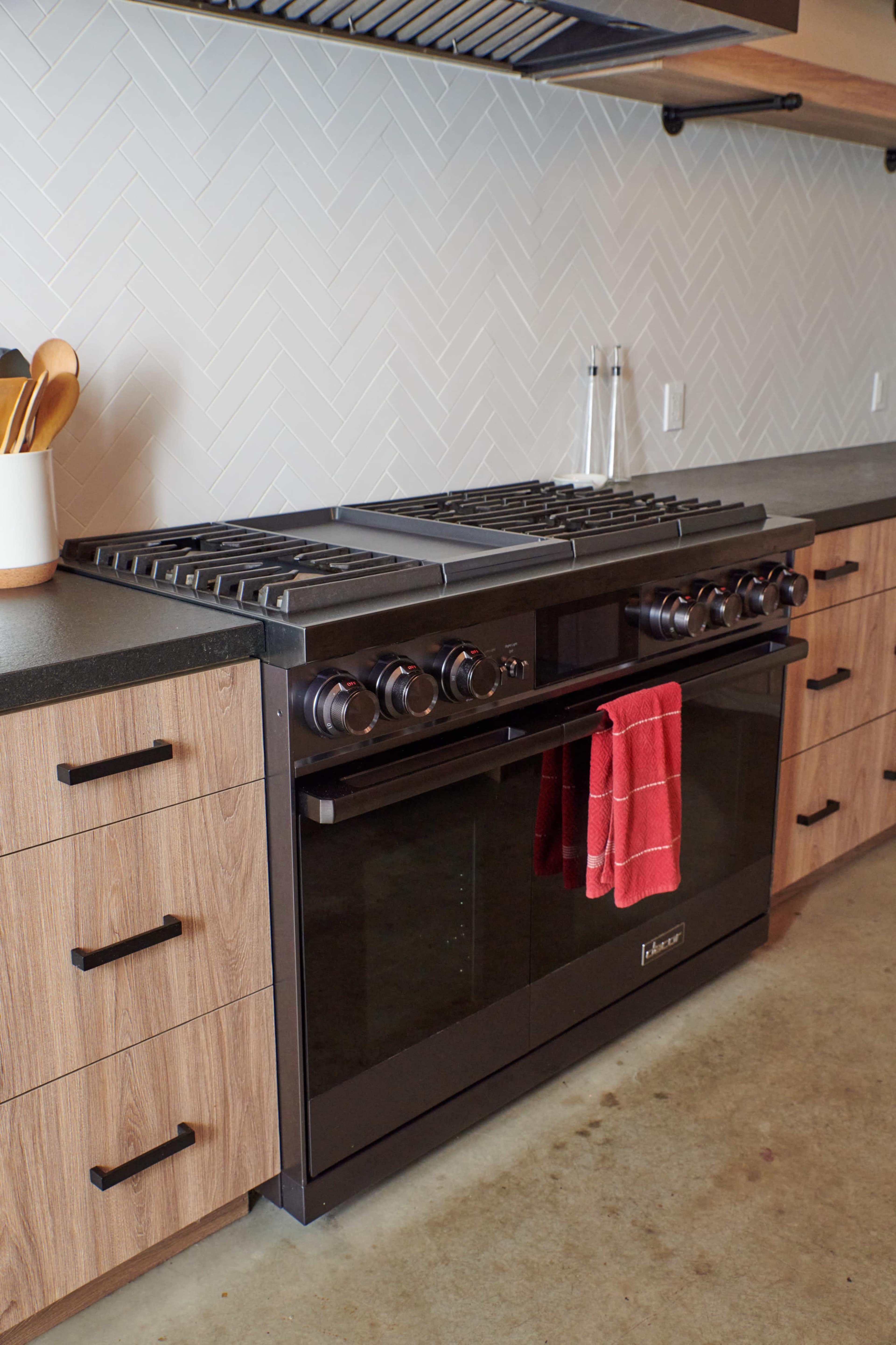 The image shows a modern kitchen with a black gas range, a red dish towel hanging from the oven handle, and wooden cabinetry beneath a herringbone-patterned backsplash.