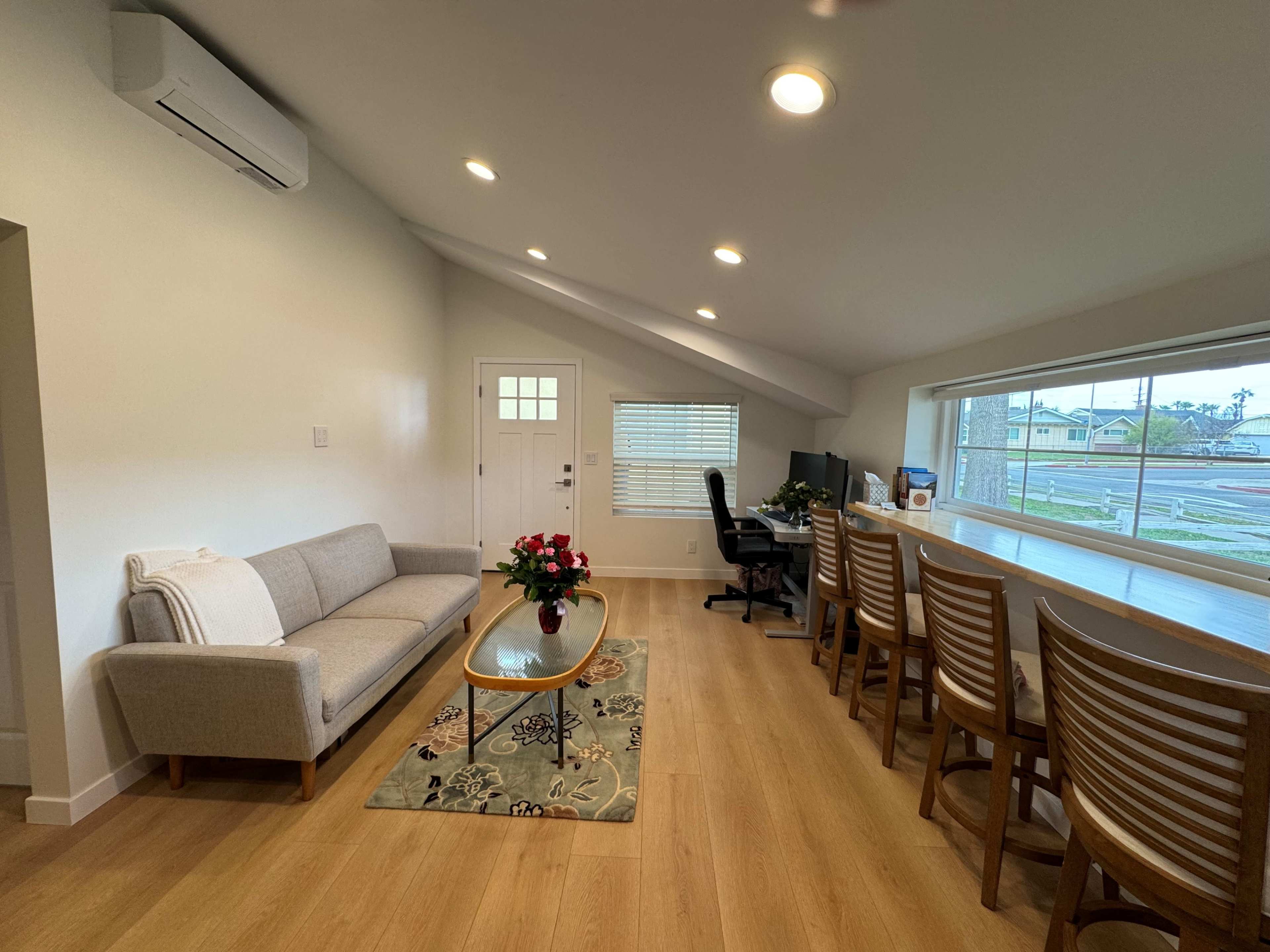 The image shows a modern living space with a gray sofa, a glass coffee table, and a long dining area with multiple wooden chairs, illuminated by ceiling lights.