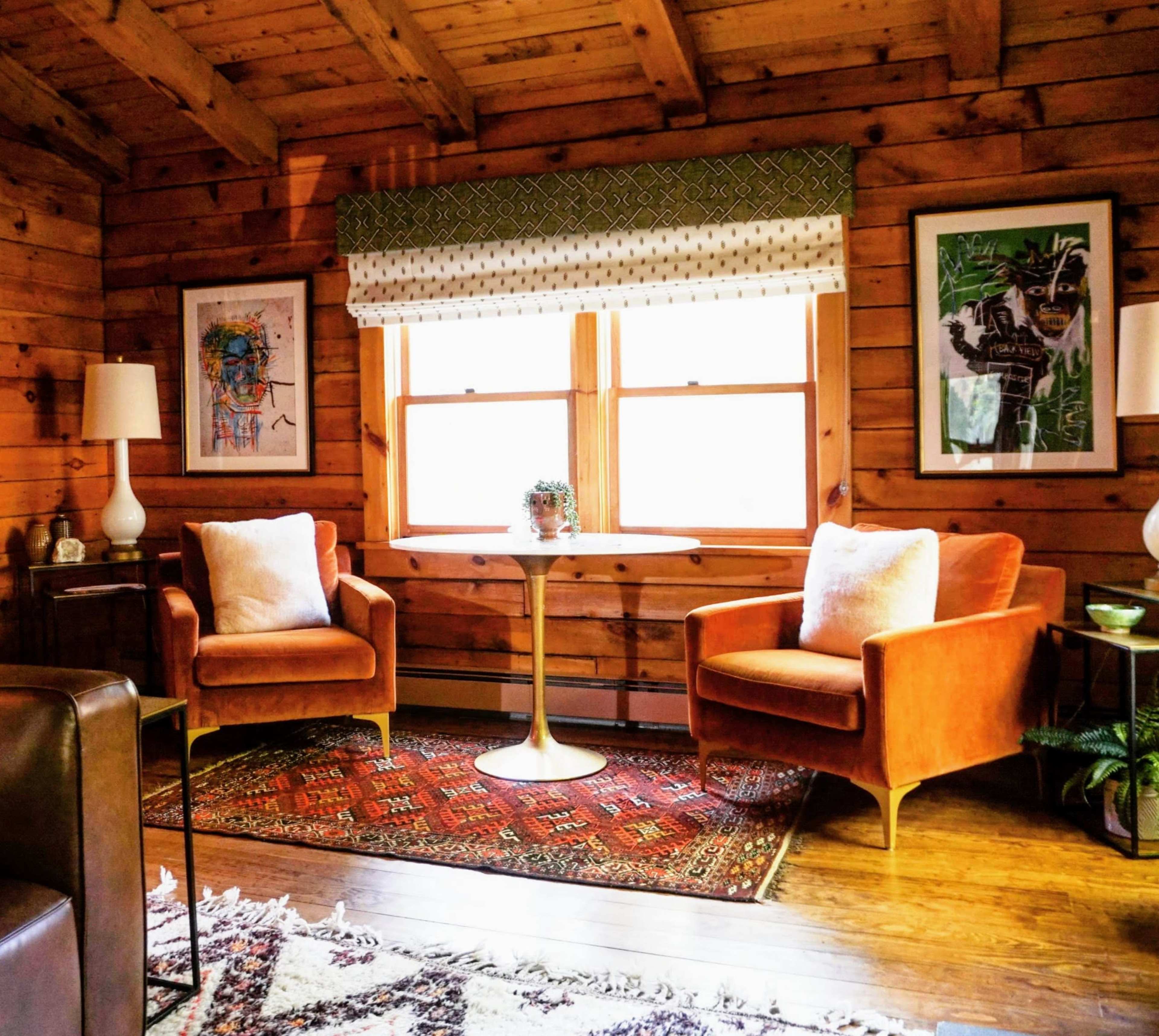 A cozy living room features two orange chairs, a round table, and large windows adorned with a patterned valance, surrounded by wooden walls and decorative artwork.