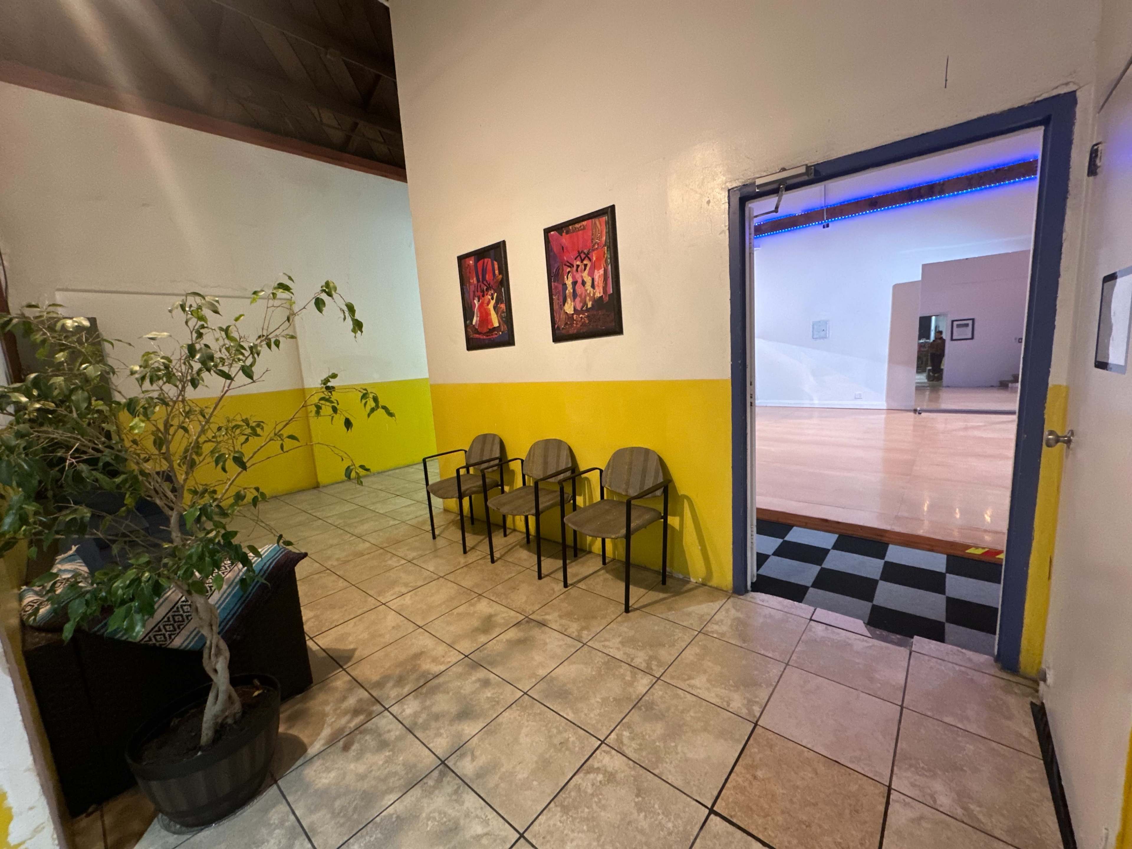 The image shows a hallway with yellow-painted lower walls, ceramic tile flooring, a small potted plant, and three chairs facing framed artwork, leading to a doorway with a visible wooden floor on the other side.