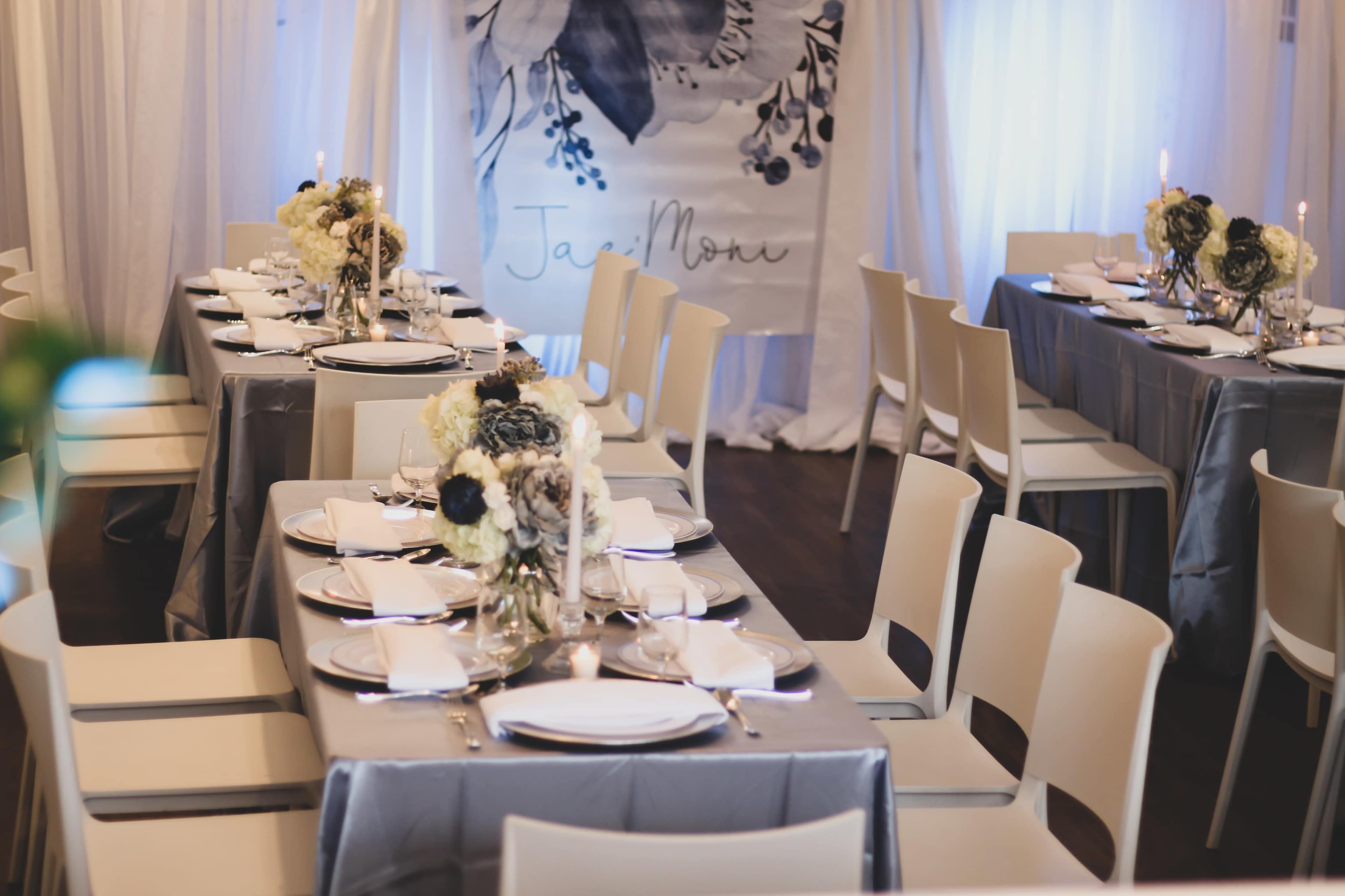 A dining setup with multiple tables covered in gray tablecloths, adorned with floral centerpieces and set with plates and candles.