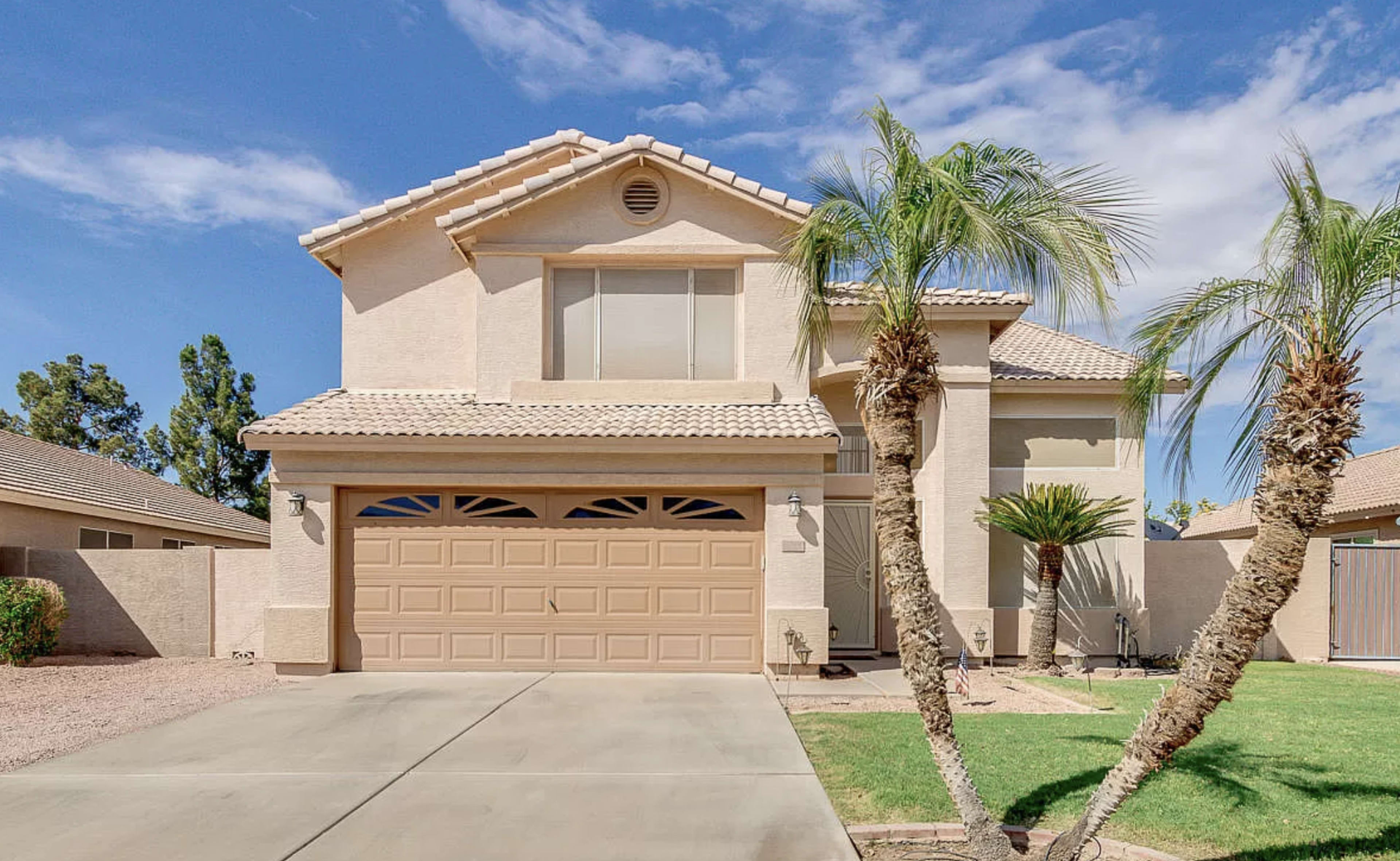 A two-story beige house with a tiled roof, palm trees in the front yard, and a two-car garage is situated under a blue sky.