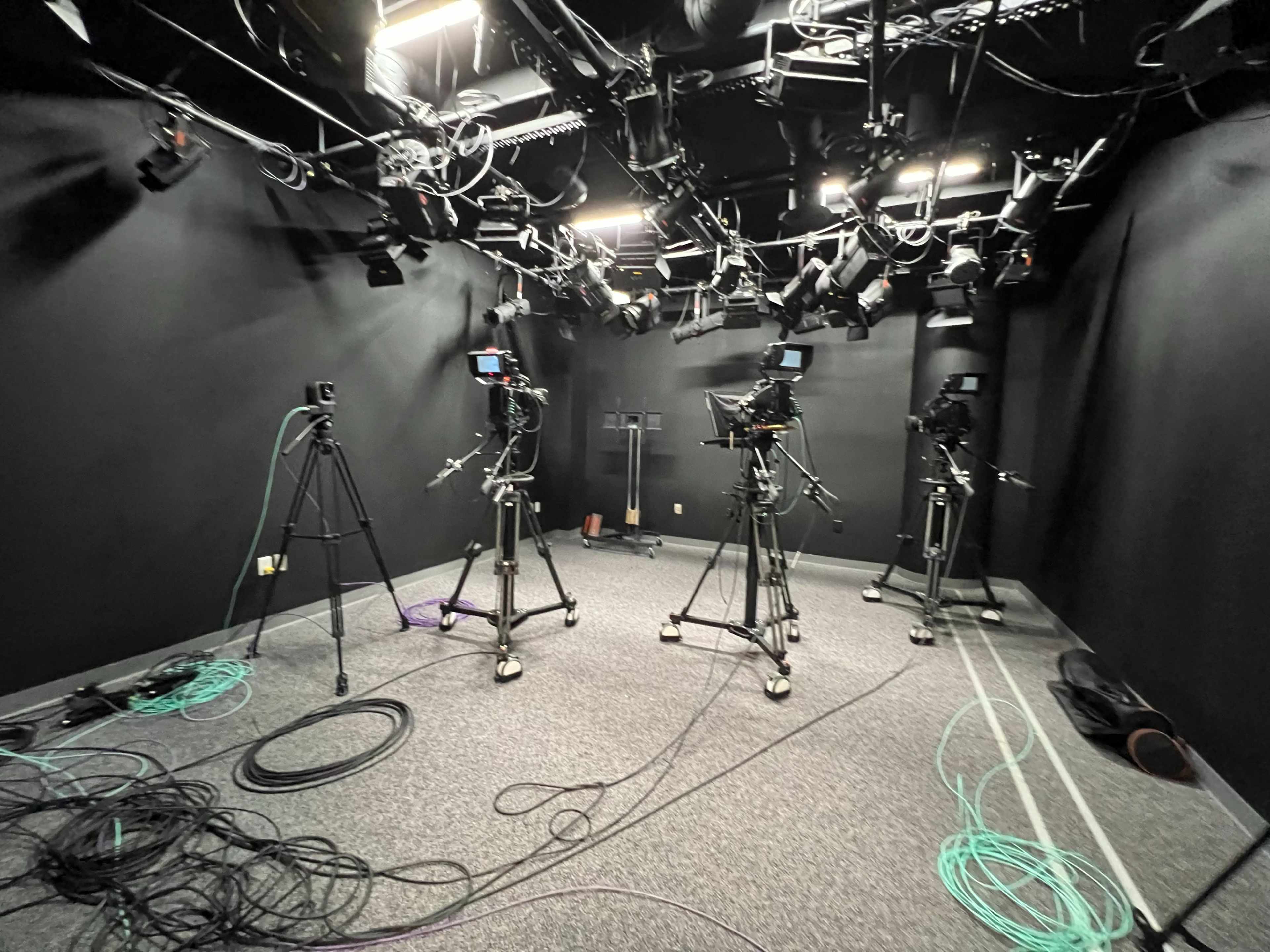 The image shows a dimly lit studio equipped with three camera setups on tripods, surrounded by various cables on the floor.
