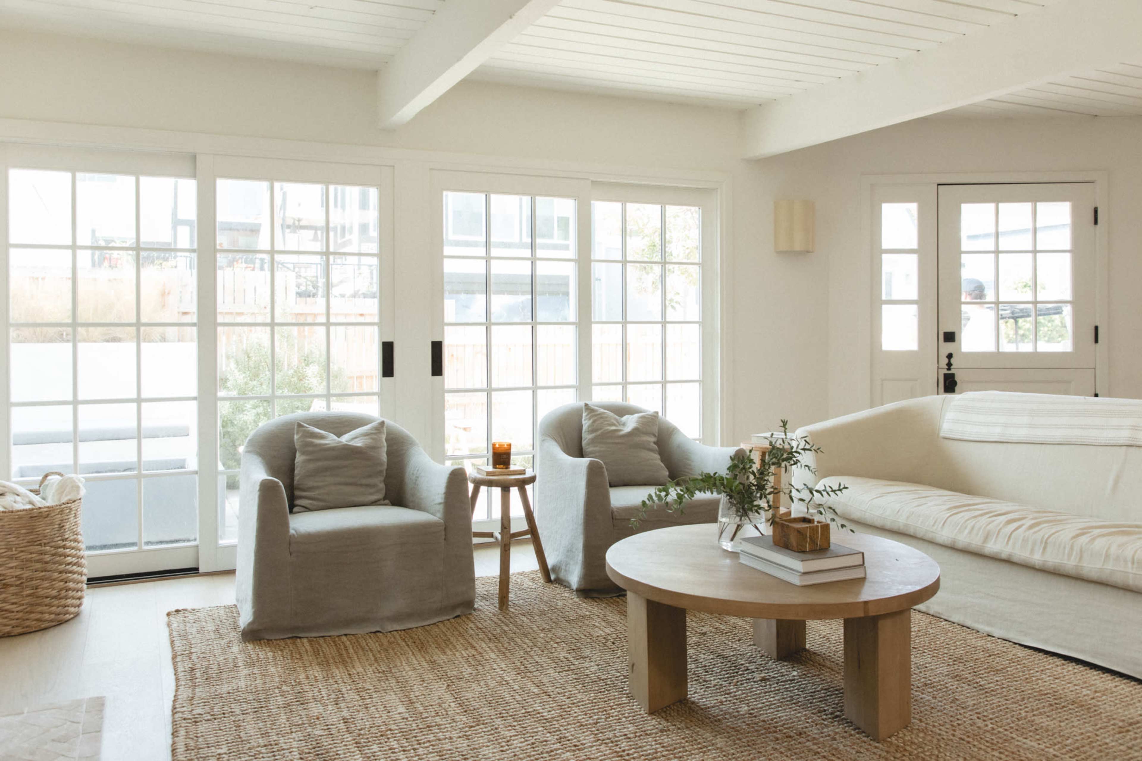The image shows a bright, modern living room with two upholstered armchairs, a round coffee table, and large windows that allow natural light to enter.