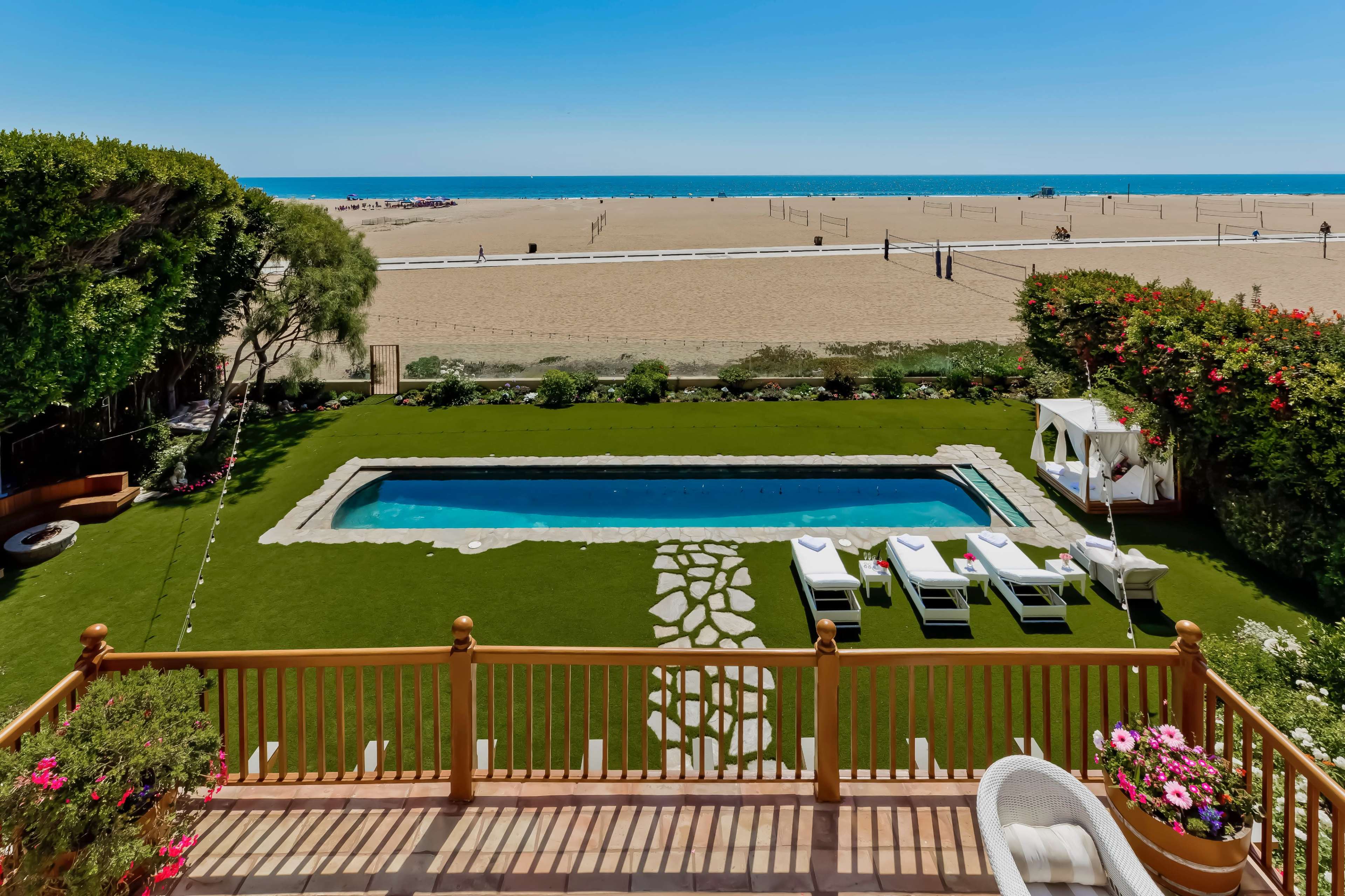The image shows a backyard with a swimming pool, lounge chairs, and a landscaped area overlooking a sandy beach and the ocean in the distance.