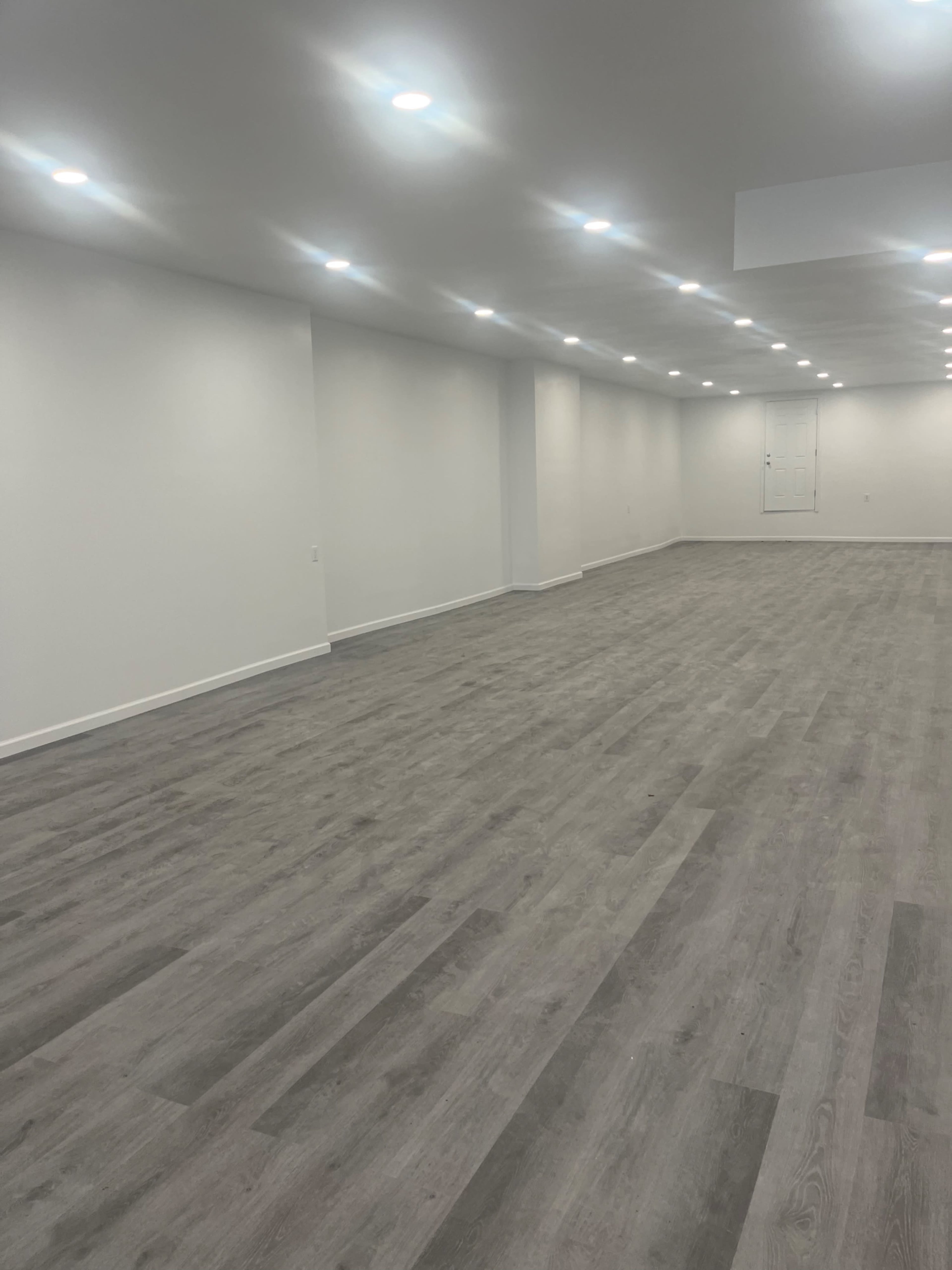 The image shows a spacious, empty room with grey hardwood flooring and ceiling lights illuminating the walls.