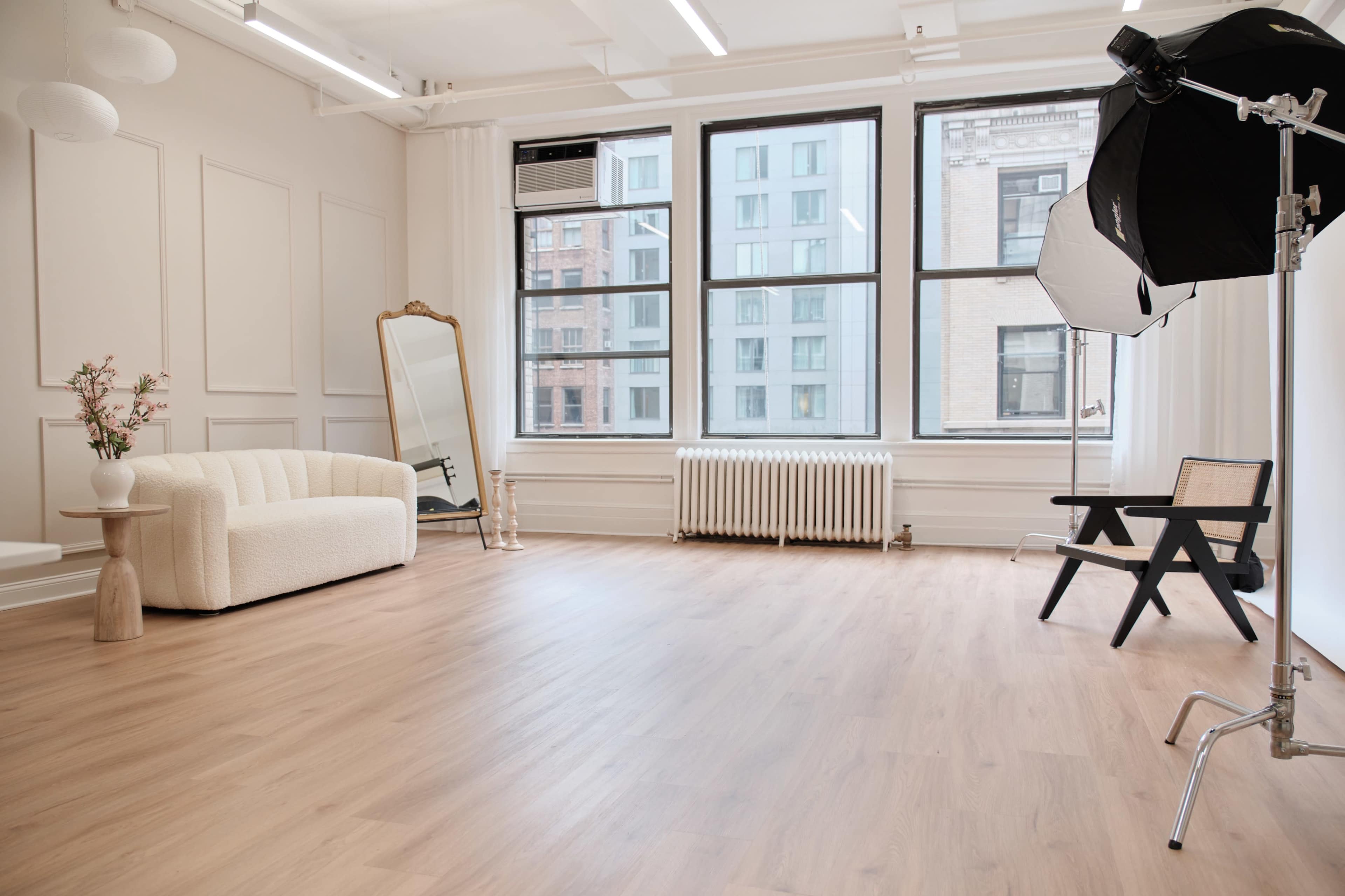 A bright, spacious studio with a white sofa, a large mirror, and natural light streaming through big windows.