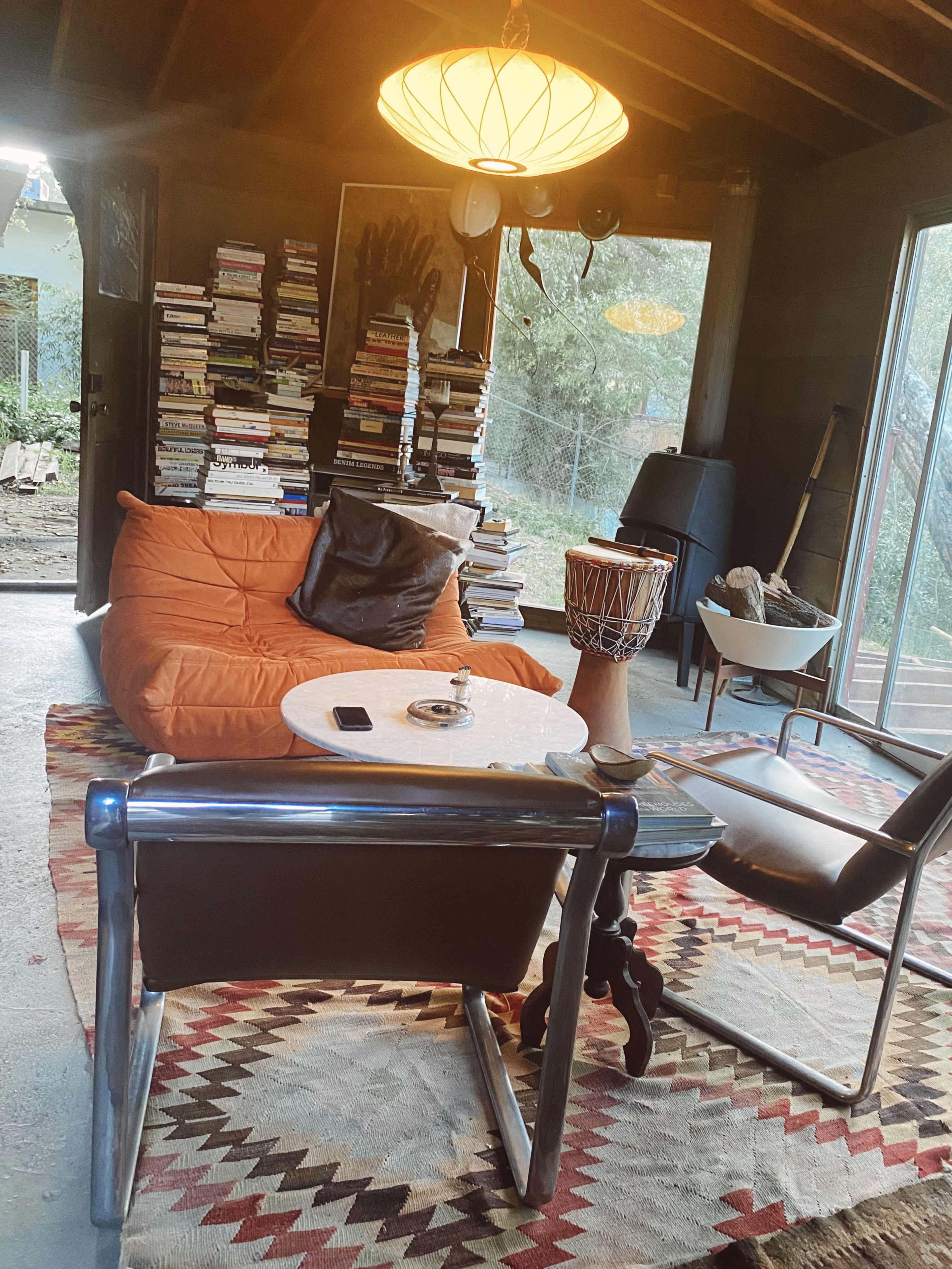 The scene features a cozy living space with an orange bean bag chair, a wood coffee table, a stack of books against the wall, and a large window overlooking a yard.