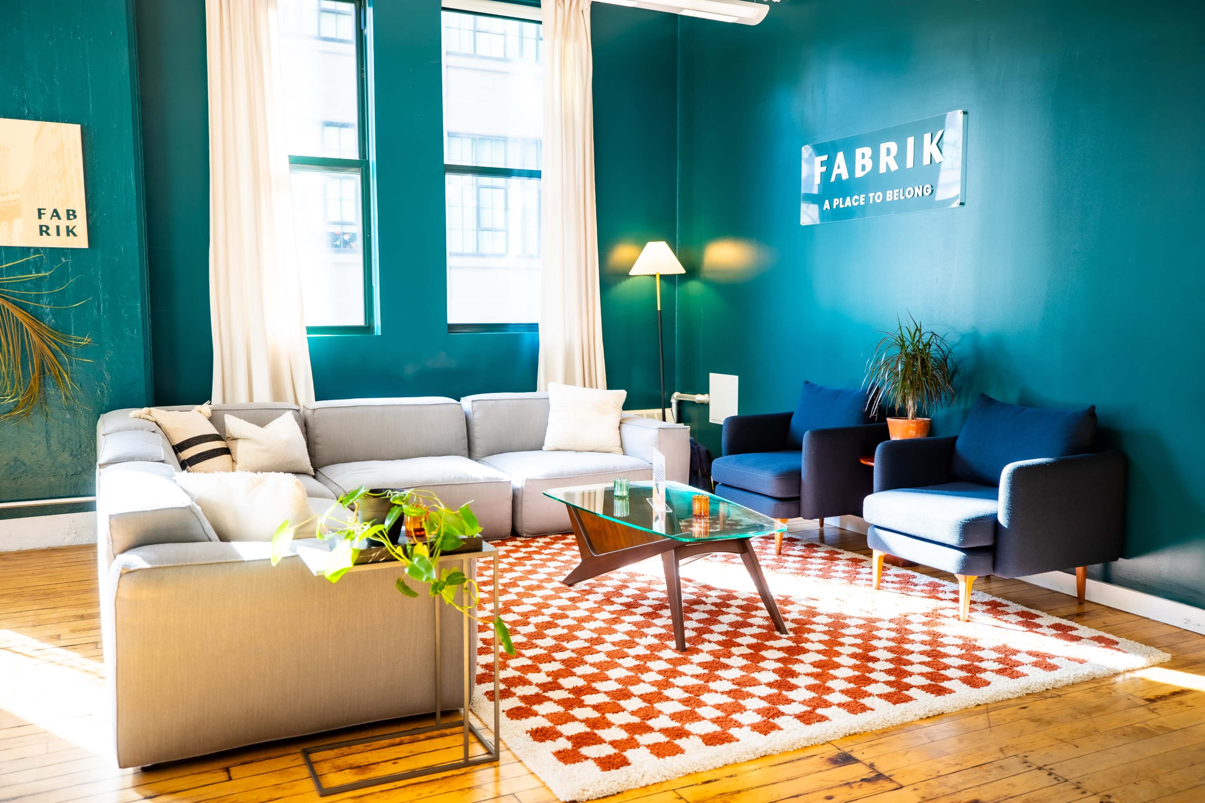 The image shows a modern lounge area featuring a gray sectional sofa, two blue chairs, a glass coffee table, and a patterned rug on a wooden floor, with green walls and large windows.