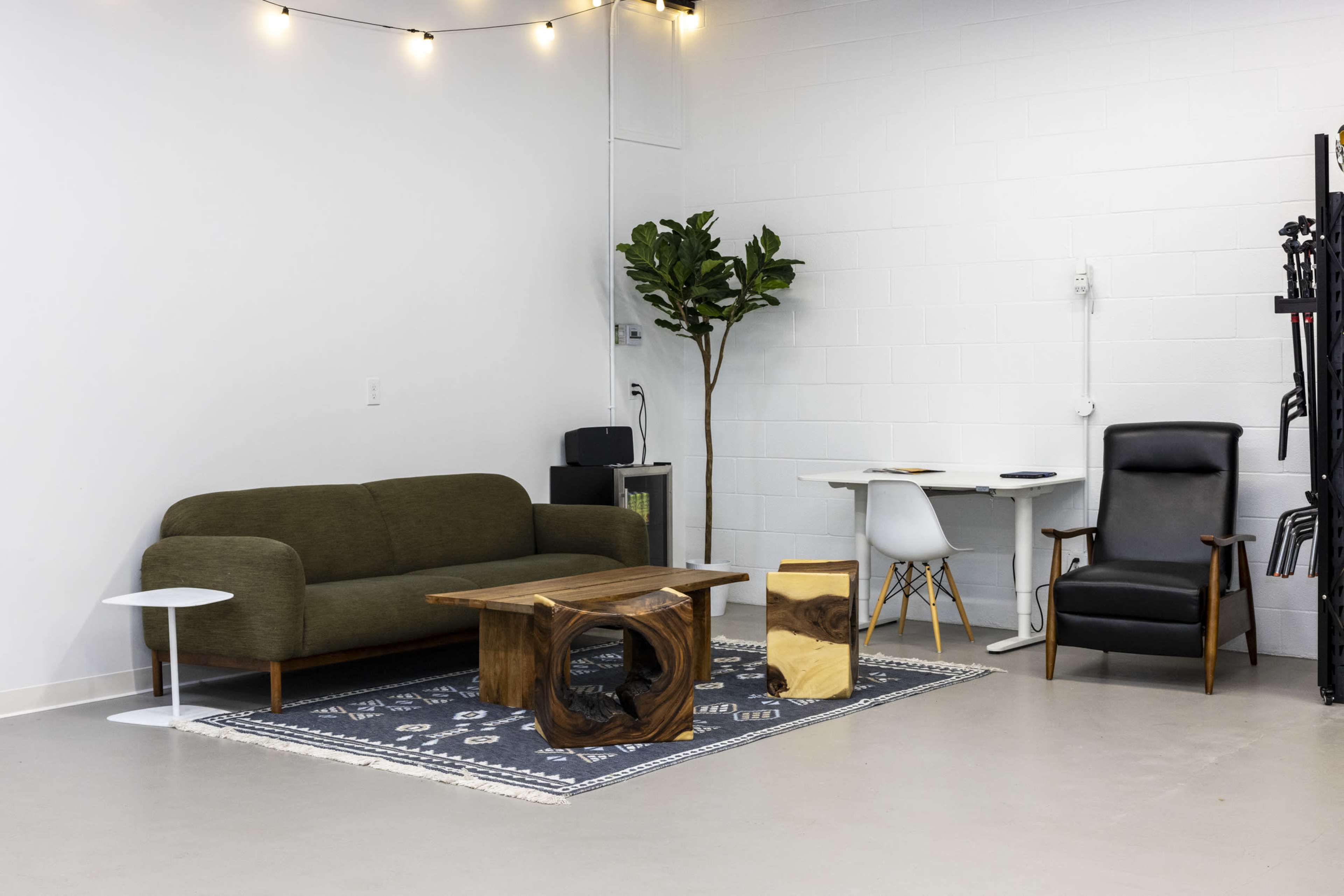 The image shows a minimalist interior design of a room featuring a green sofa, a wooden coffee table, a potted plant, a desk with a white chair, and a black armchair on a patterned rug.