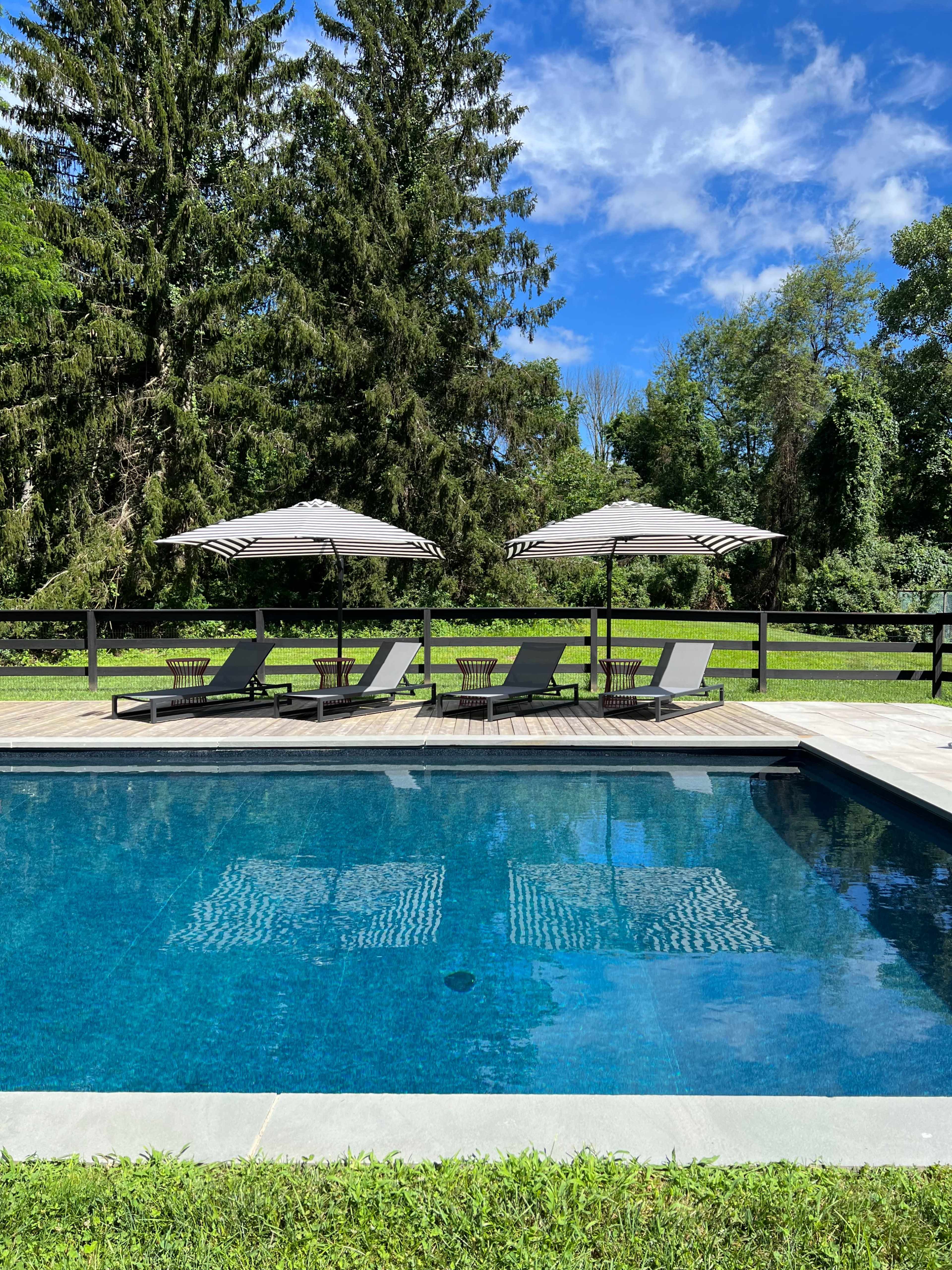 A serene outdoor pool area features lounge chairs under large umbrellas, surrounded by lush greenery and a clear blue sky.