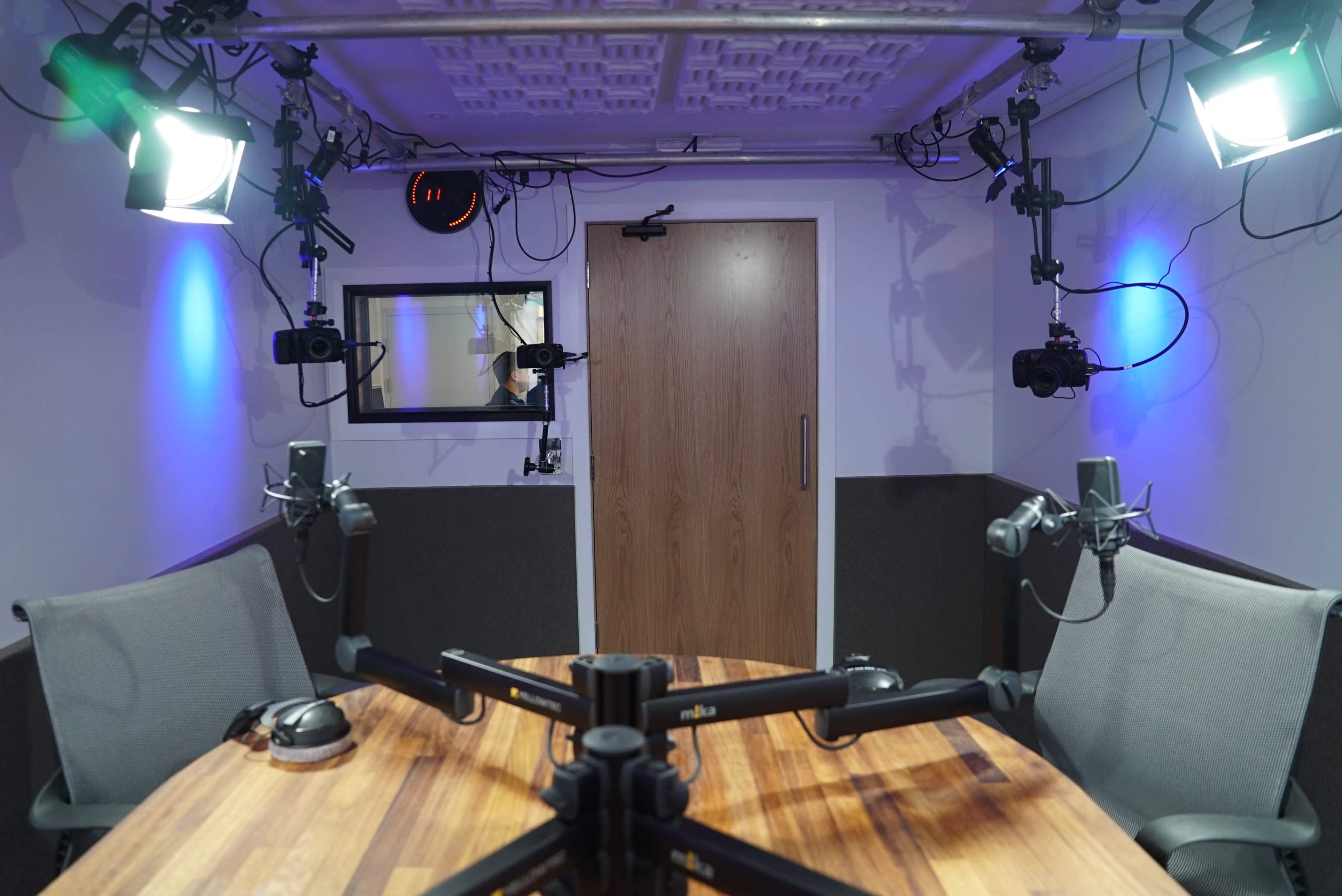 A small recording studio features a wooden table with two chairs, equipped with multiple microphones and cameras, along with professional lighting setup.