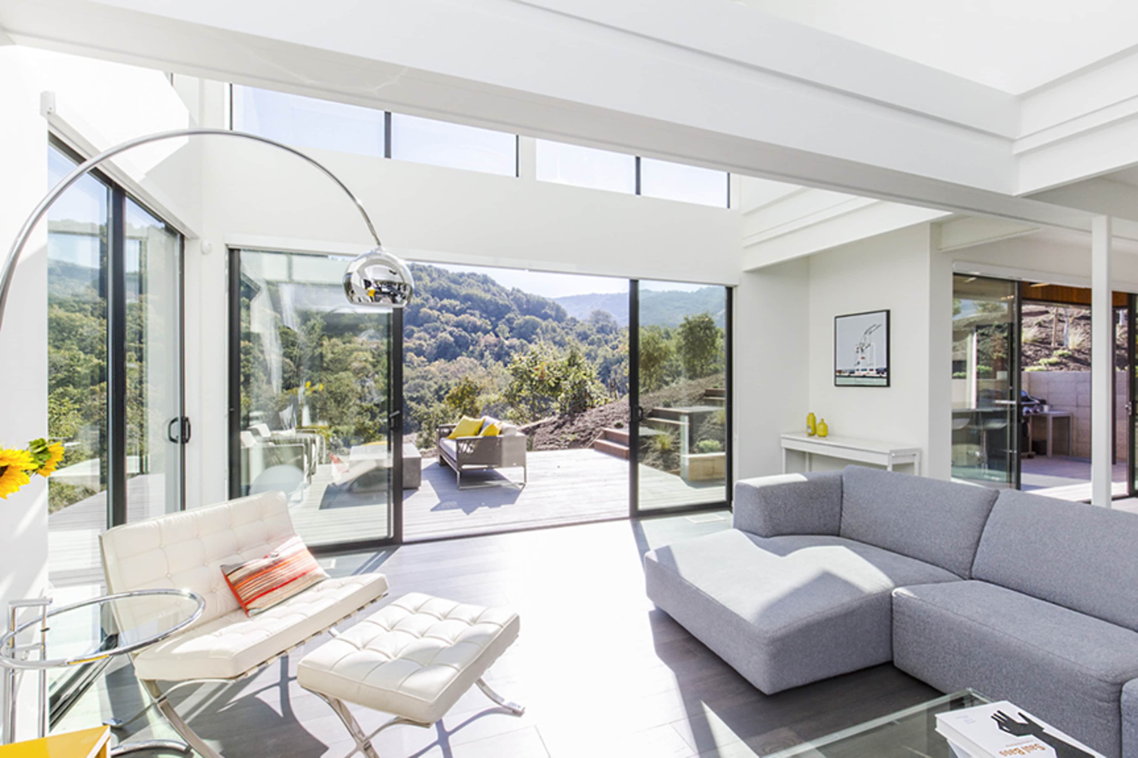 A modern living room features large glass windows that open to a view of grassy hills and outdoor seating.