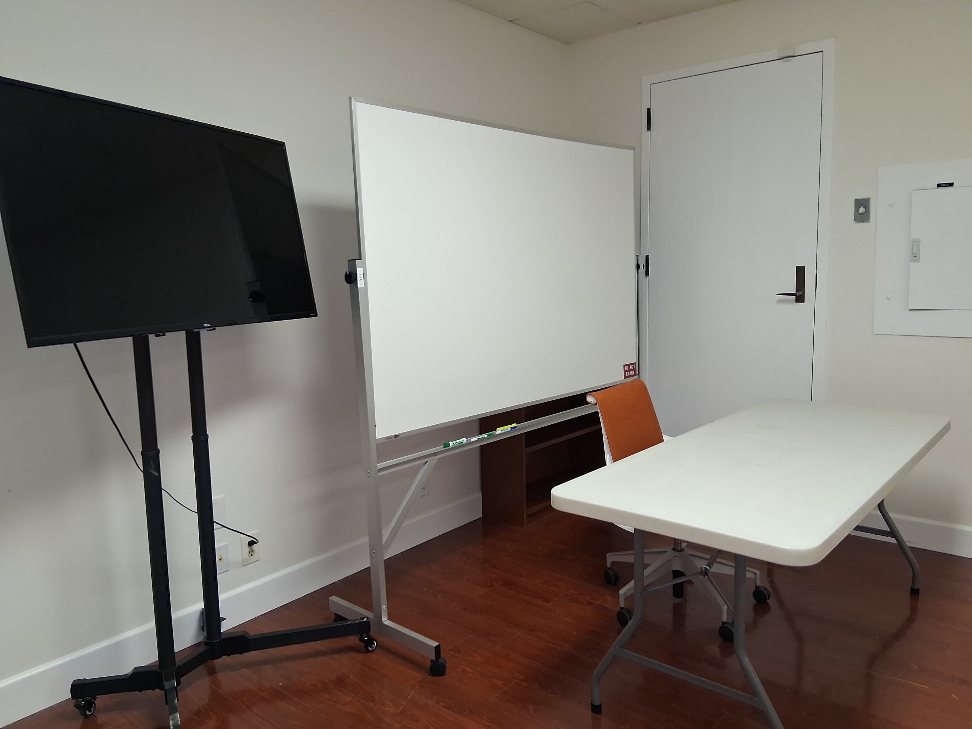 The image shows a small meeting room with a TV on a stand, a whiteboard on wheels, and a table with a chair.