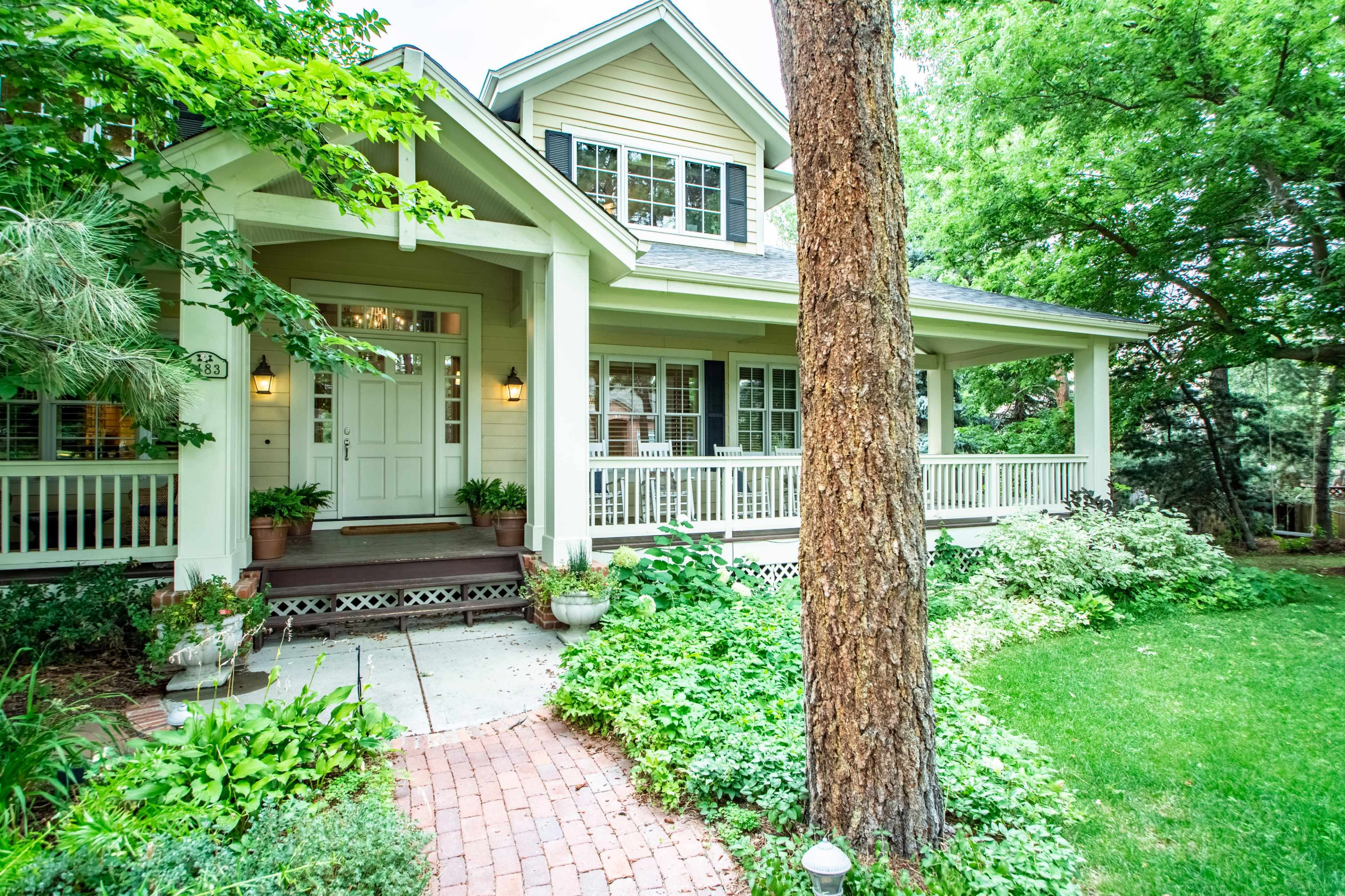 A two-story house with a large front porch, surrounded by greenery and brick pathways.