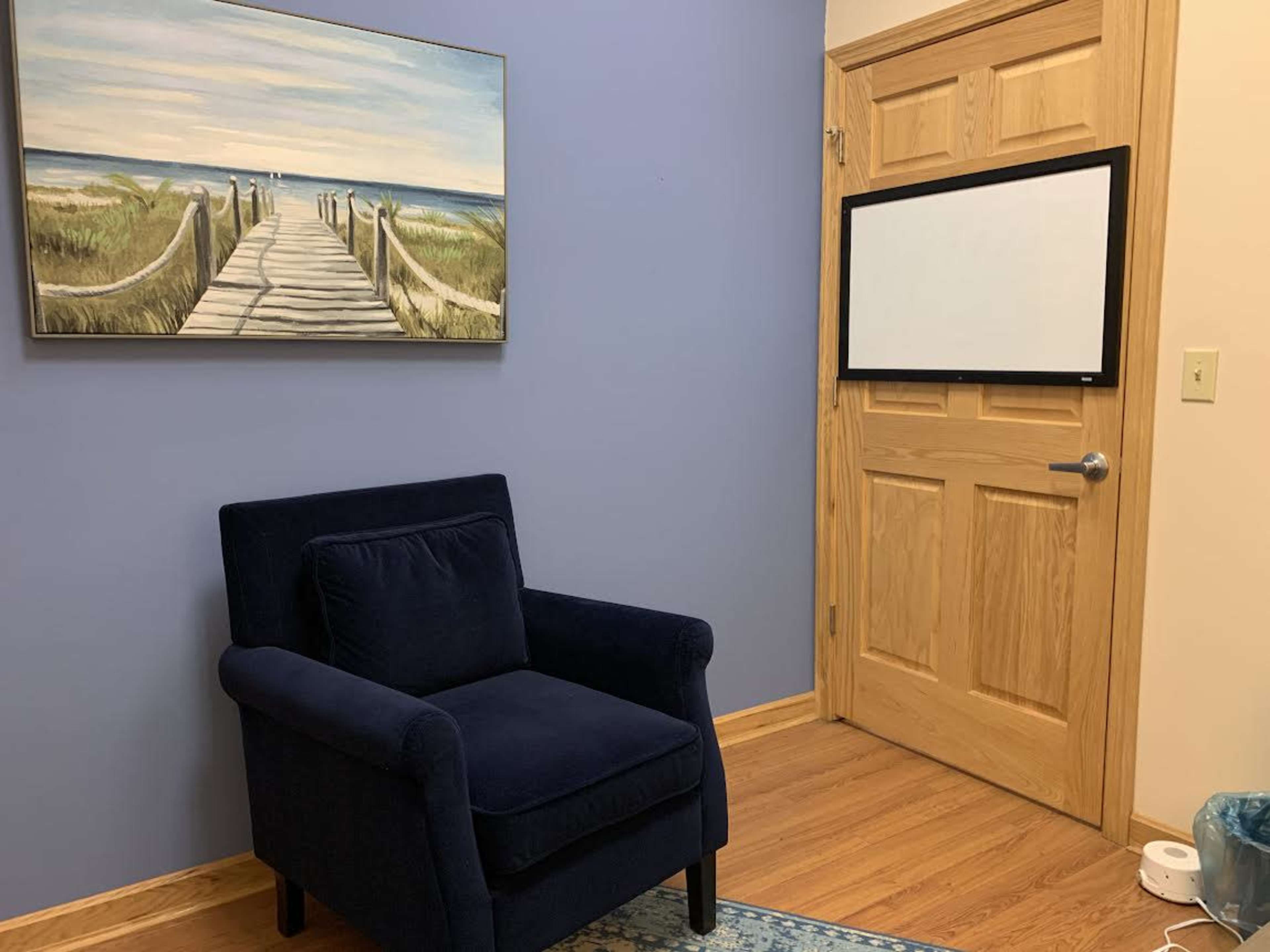 The image shows a room with a blue wall, a wooden door, a painting of a boardwalk leading to the beach, a blue armchair, and a blank wall-mounted screen.