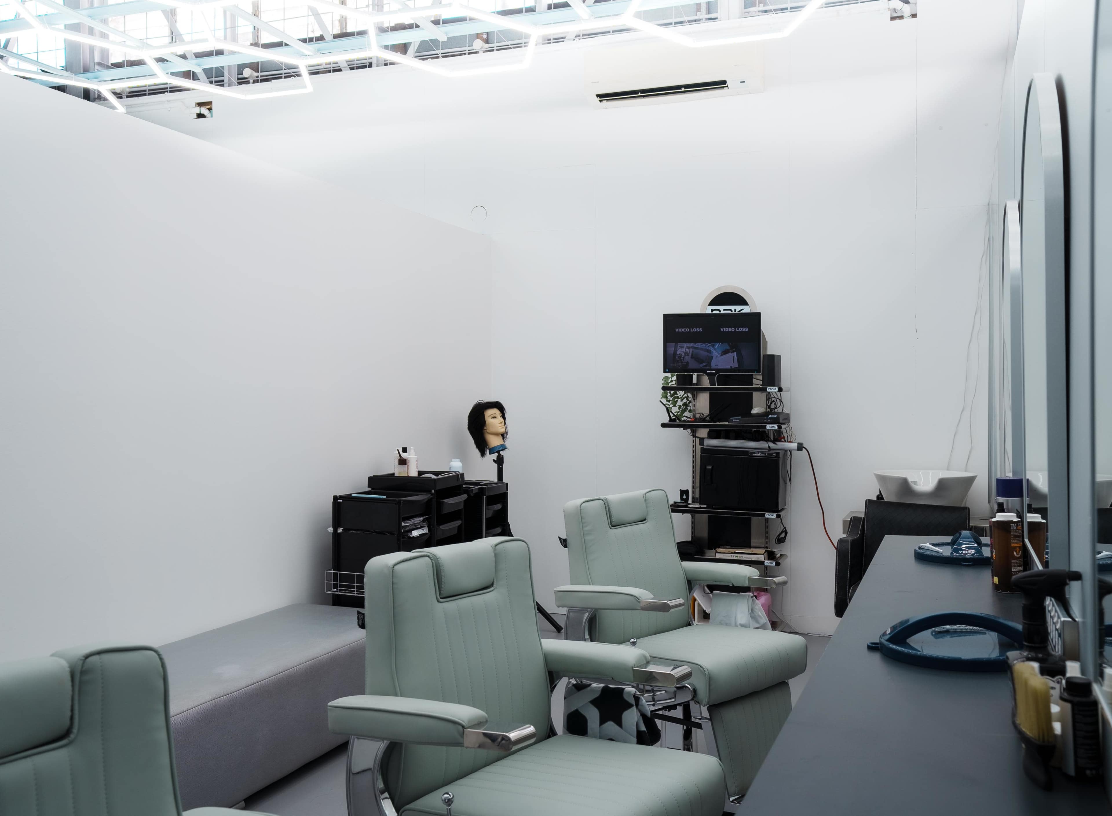 A modern barbershop features two green barber chairs, a waiting area, and a monitor on a cart, with a mannequin head displayed nearby.