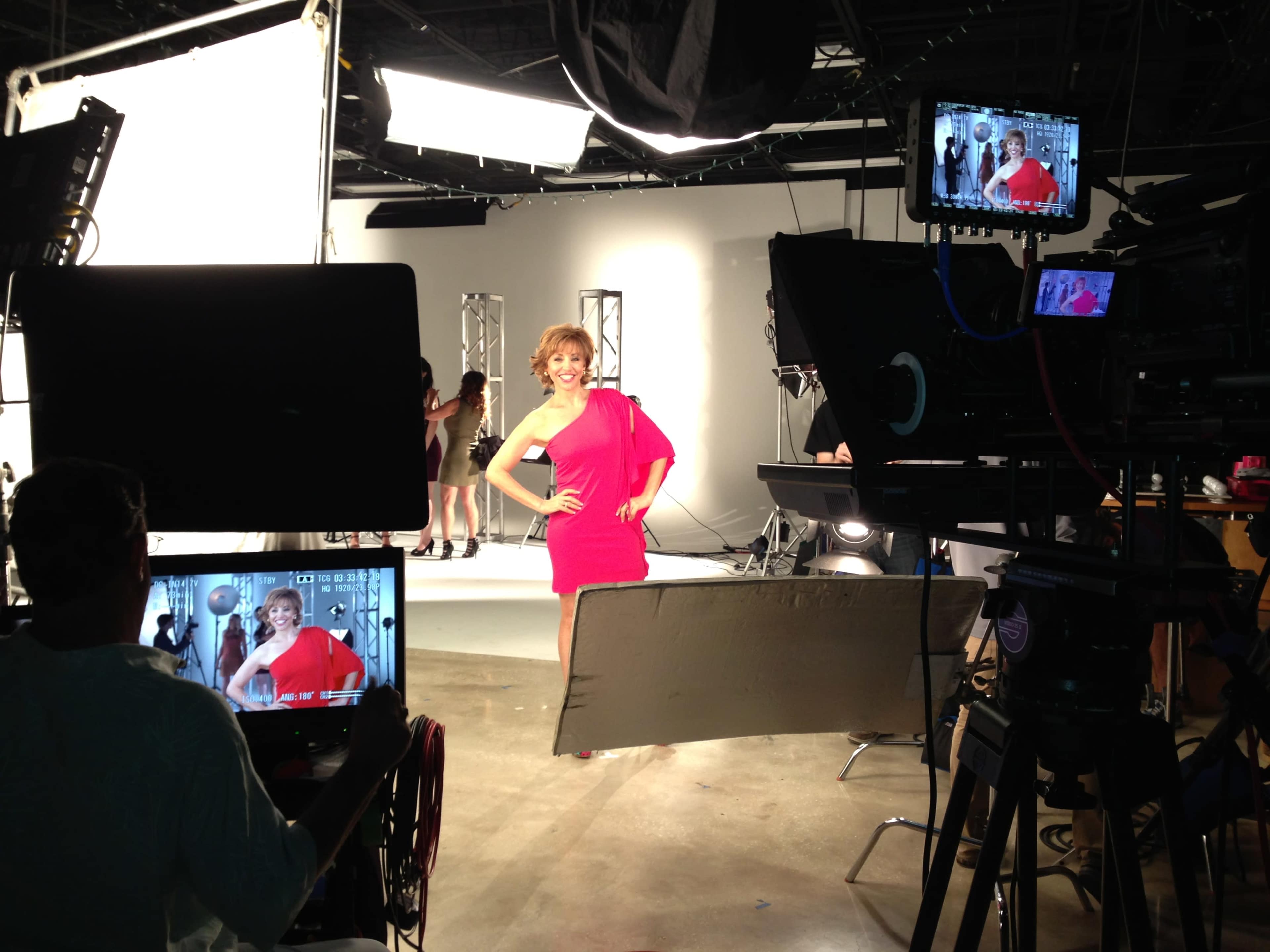 A woman in a bright pink dress poses confidently in a studio surrounded by cameras and lighting equipment.