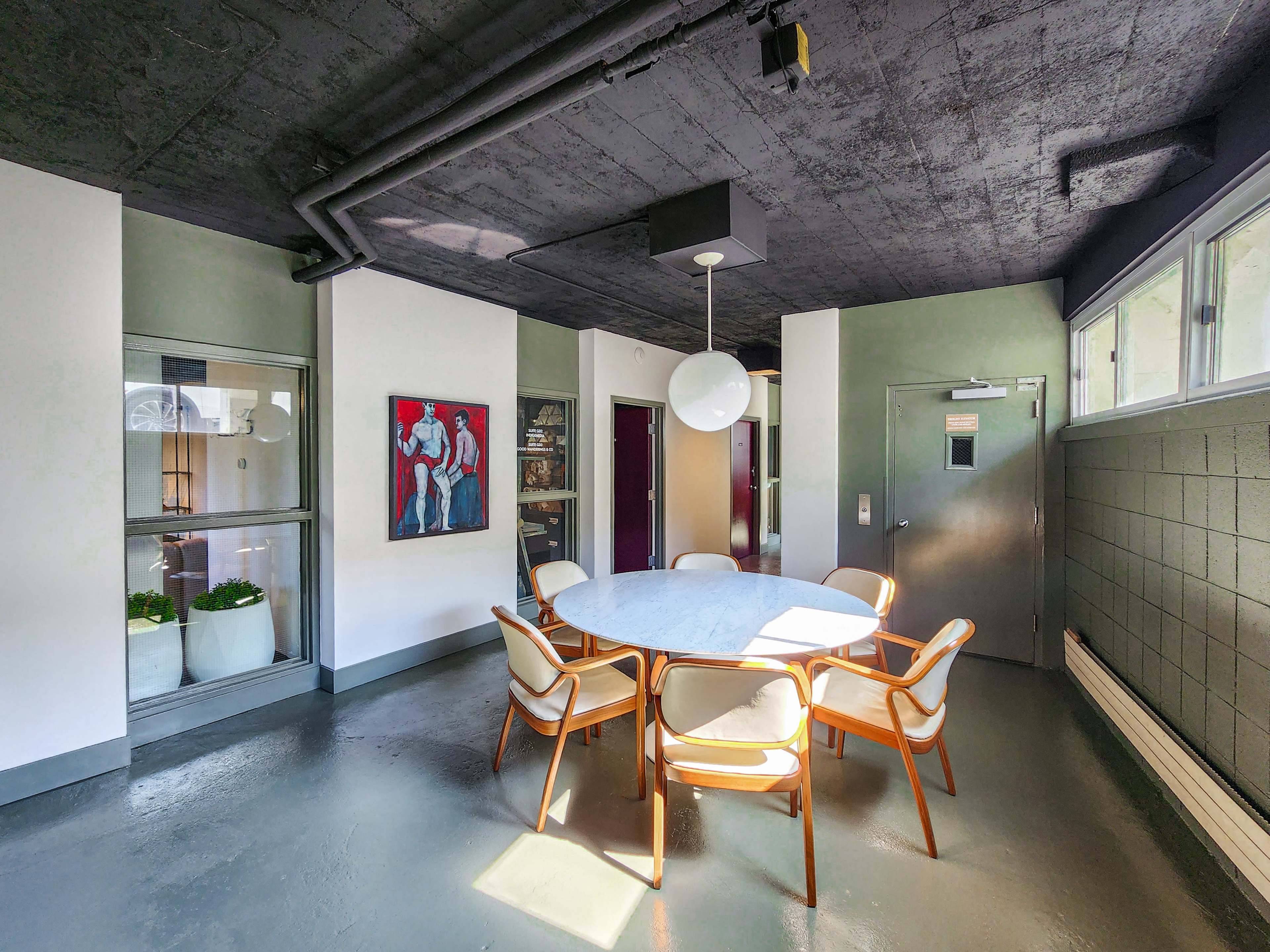 The image shows a modern interior space featuring a round dining table surrounded by white chairs, with a painting on the wall and large windows letting in natural light.