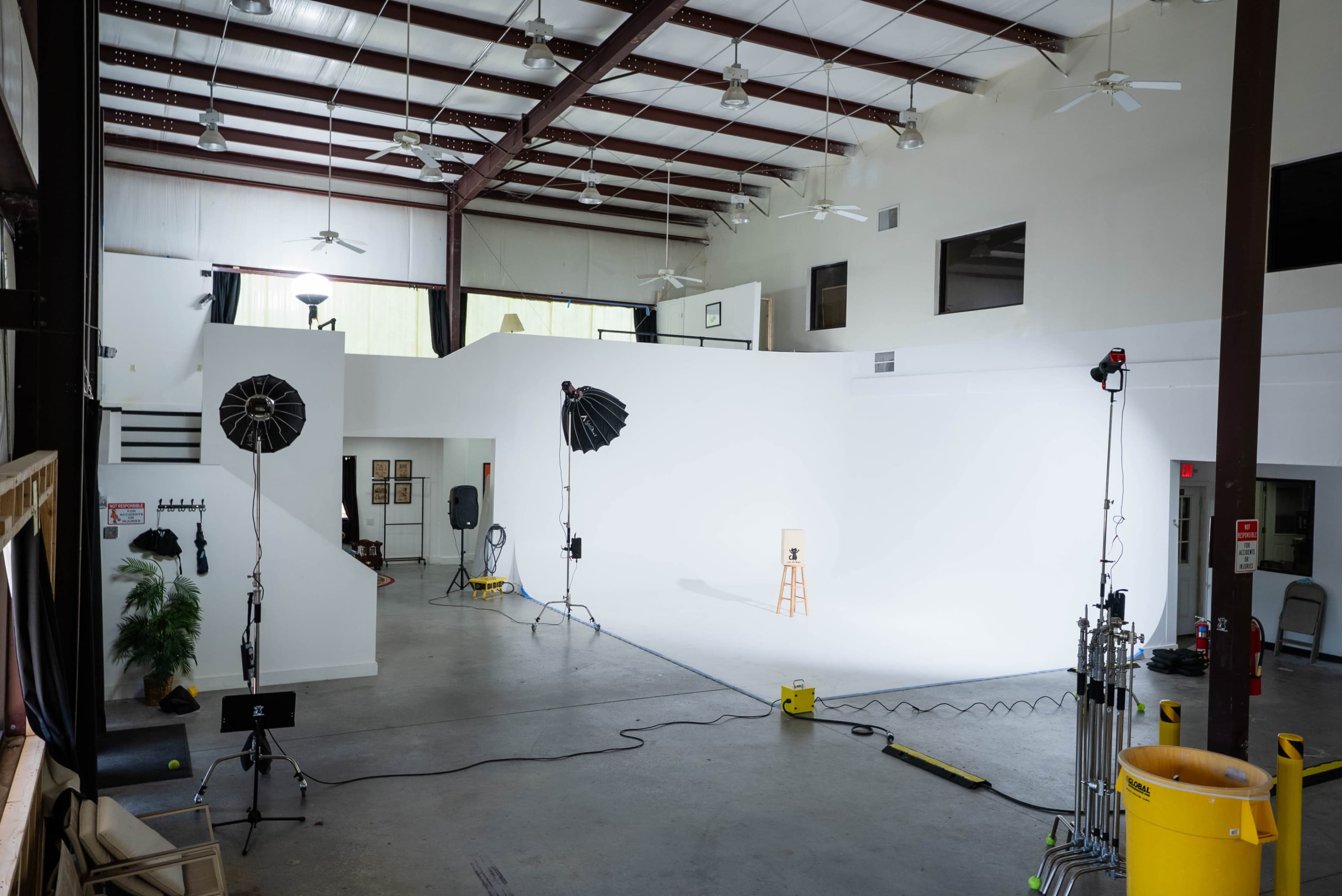 The image shows a spacious photography studio with a large white backdrop, various lighting equipment, and a wooden stool positioned in the center.