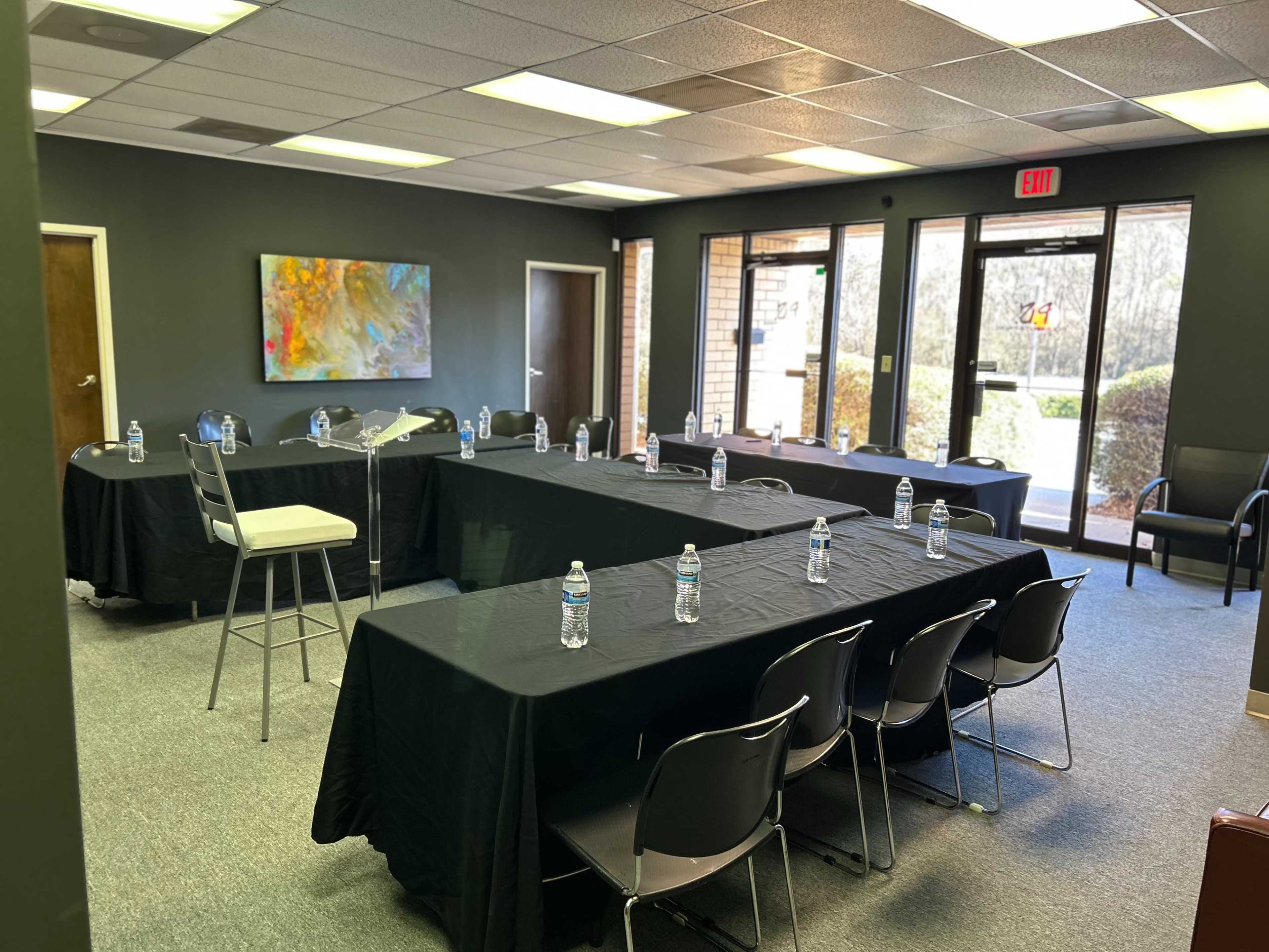 The image shows a conference room with several black-covered tables arranged in a U-shape, each equipped with a water bottle, and a large screen displaying art on the wall.