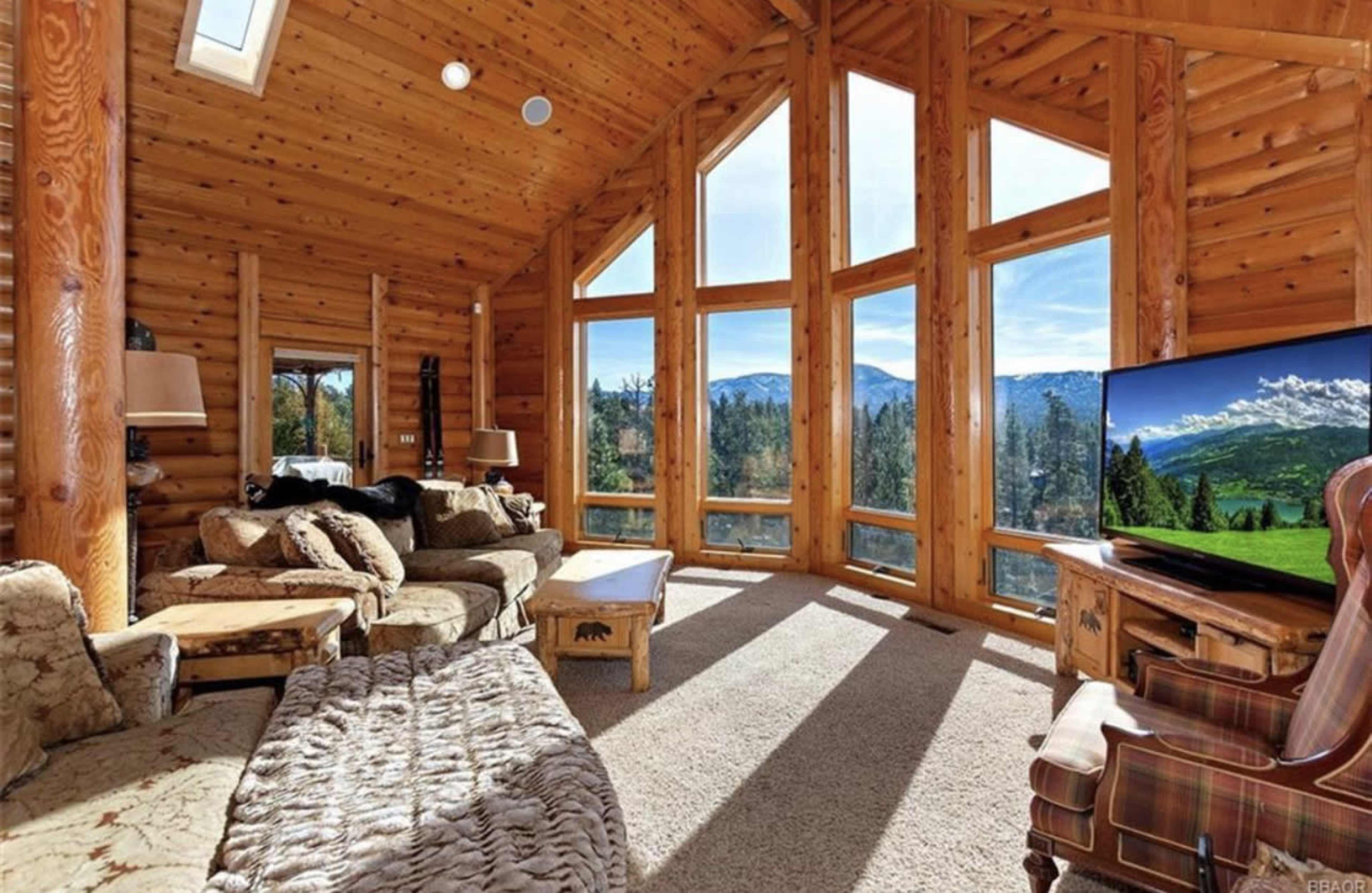 A spacious cabin interior features large windows with mountain views, a cozy seating area, and a television.