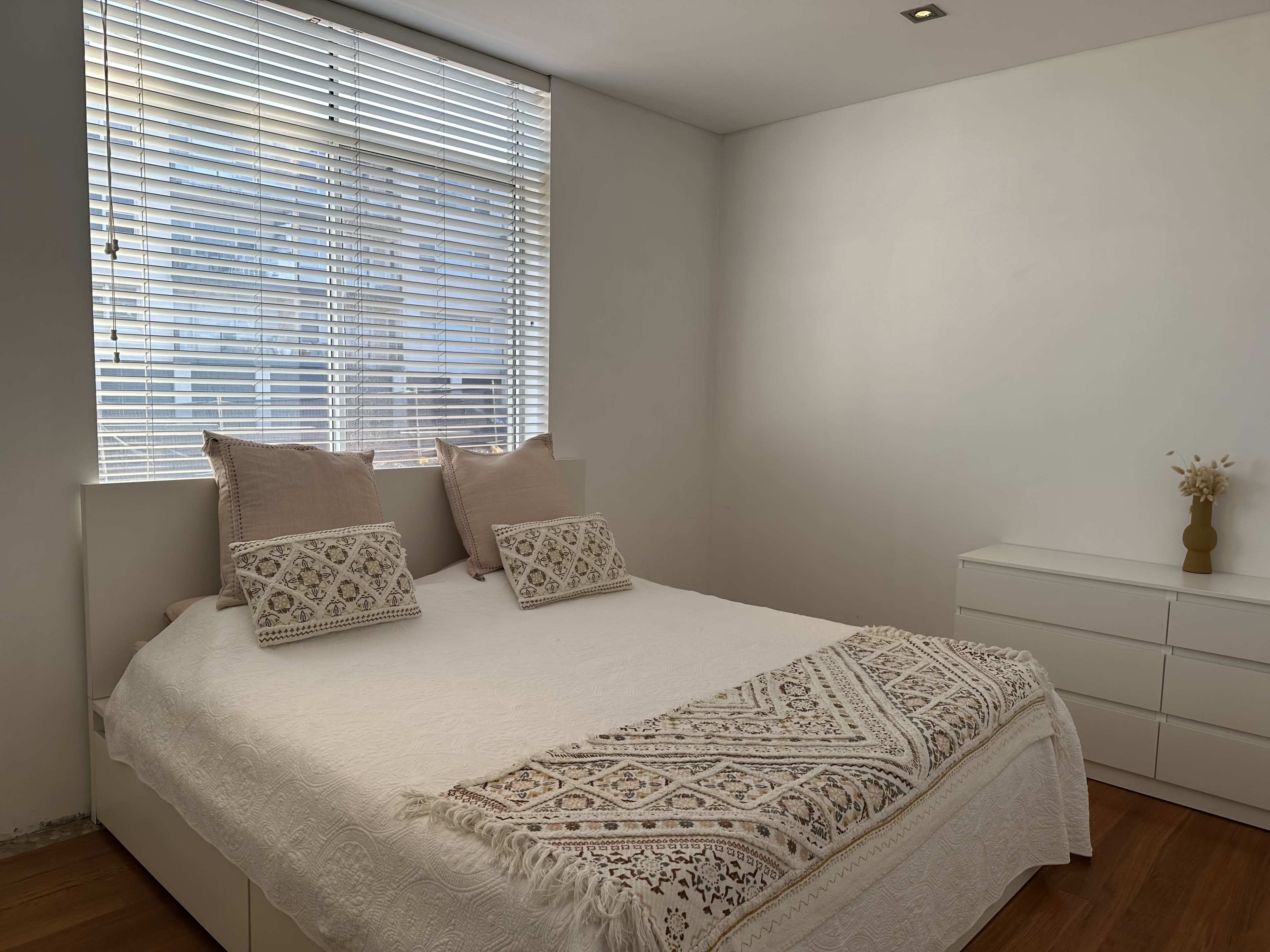 A neatly arranged bedroom with a large bed featuring two decorative pillows, a window with blinds, and a dresser against the wall.