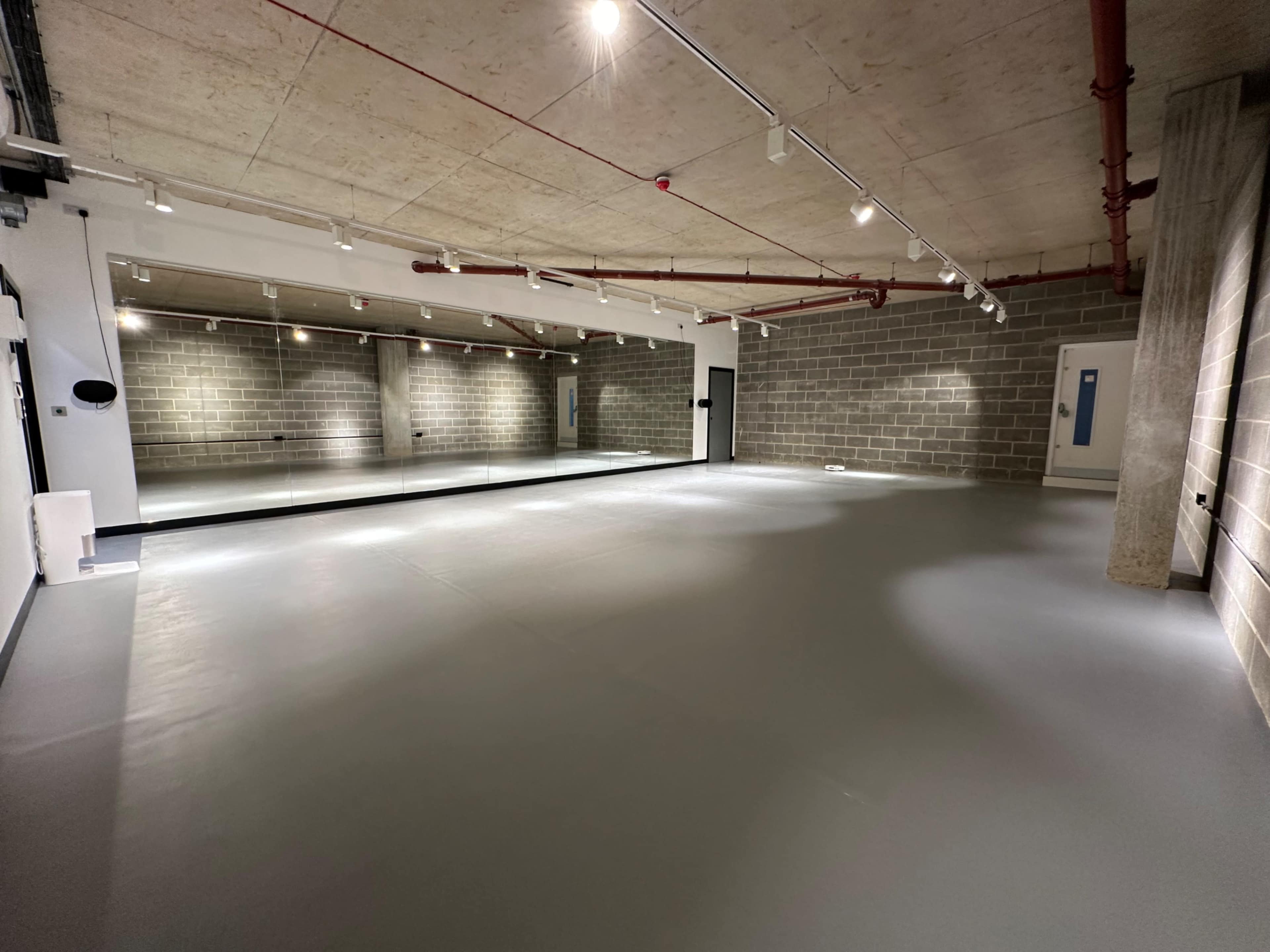 The image shows a spacious, empty room with gray flooring and a large mirror along one wall, surrounded by exposed concrete and industrial lighting.
