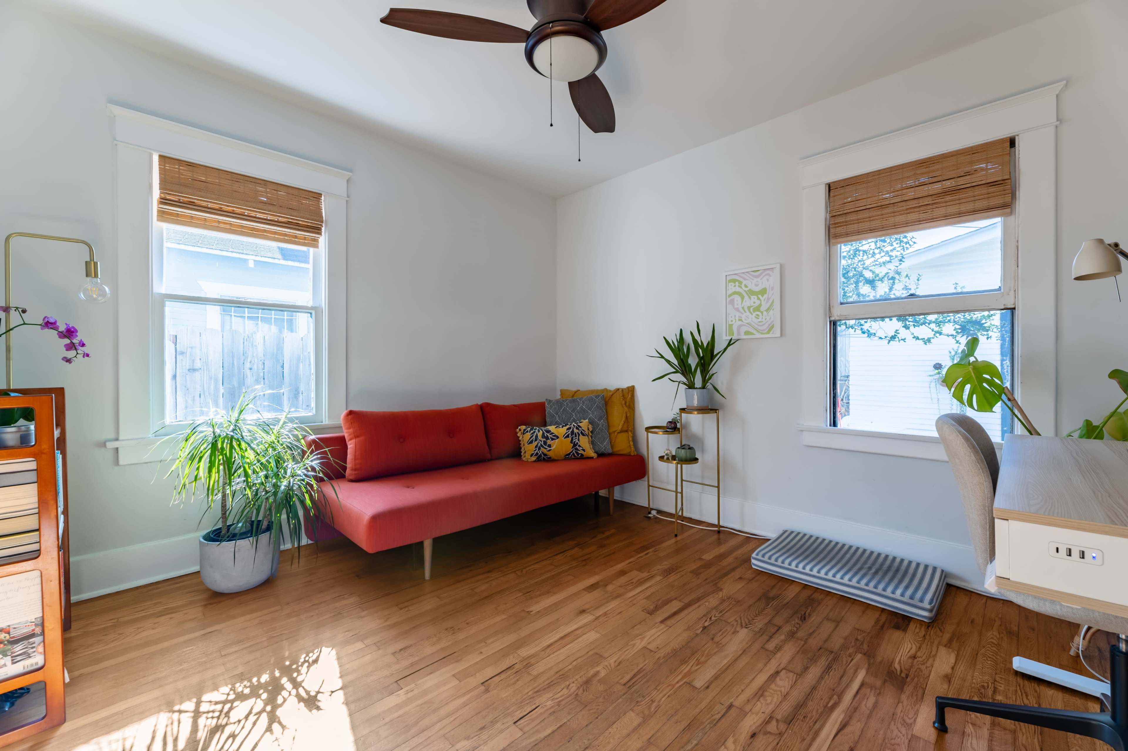 The image shows a bright room featuring a red sofa, potted plants, and a small desk with a chair, illuminated by natural light from two windows.