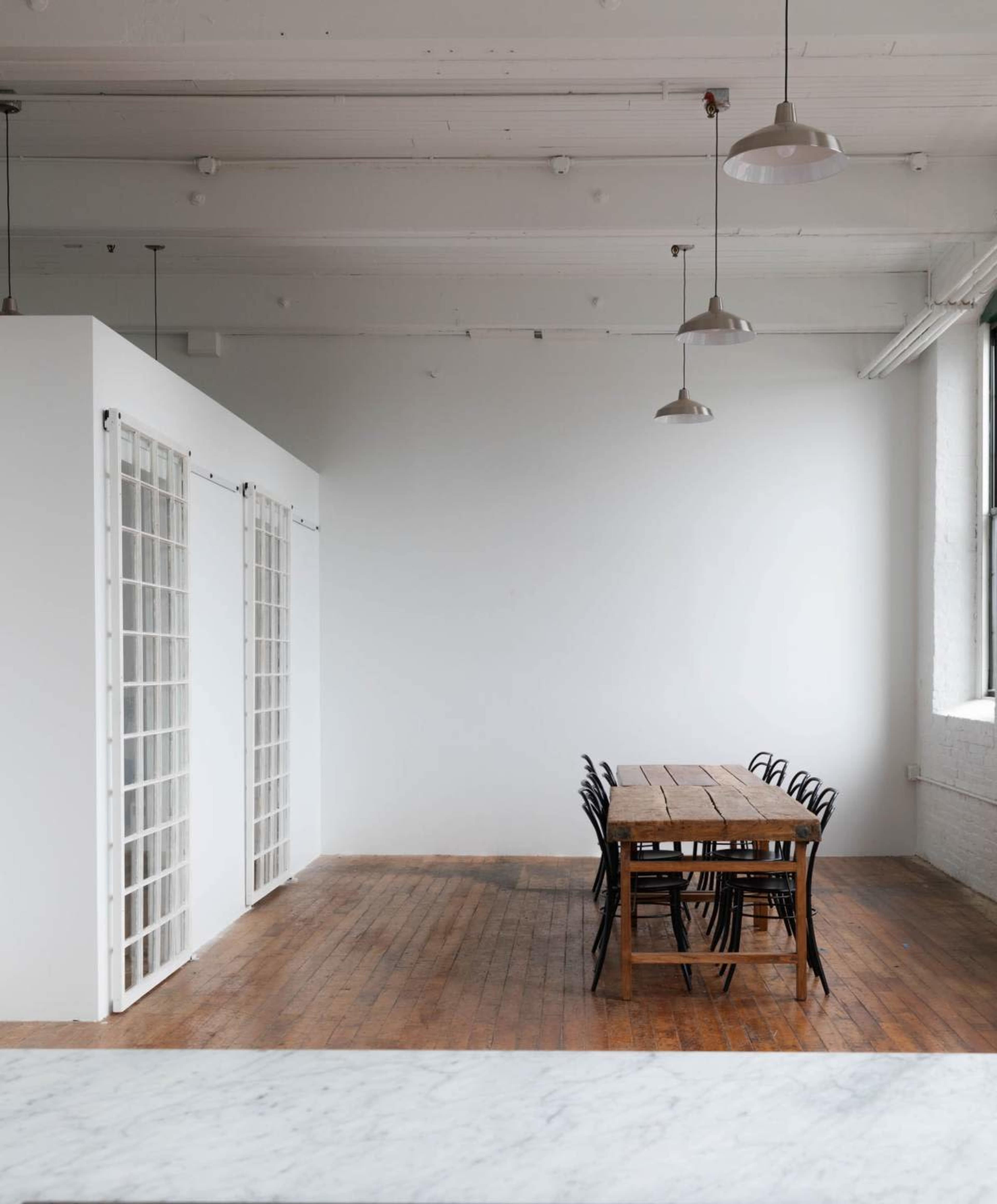A minimalistic interior space features a long wooden table surrounded by black chairs, with a bright white wall and large windows in the background.