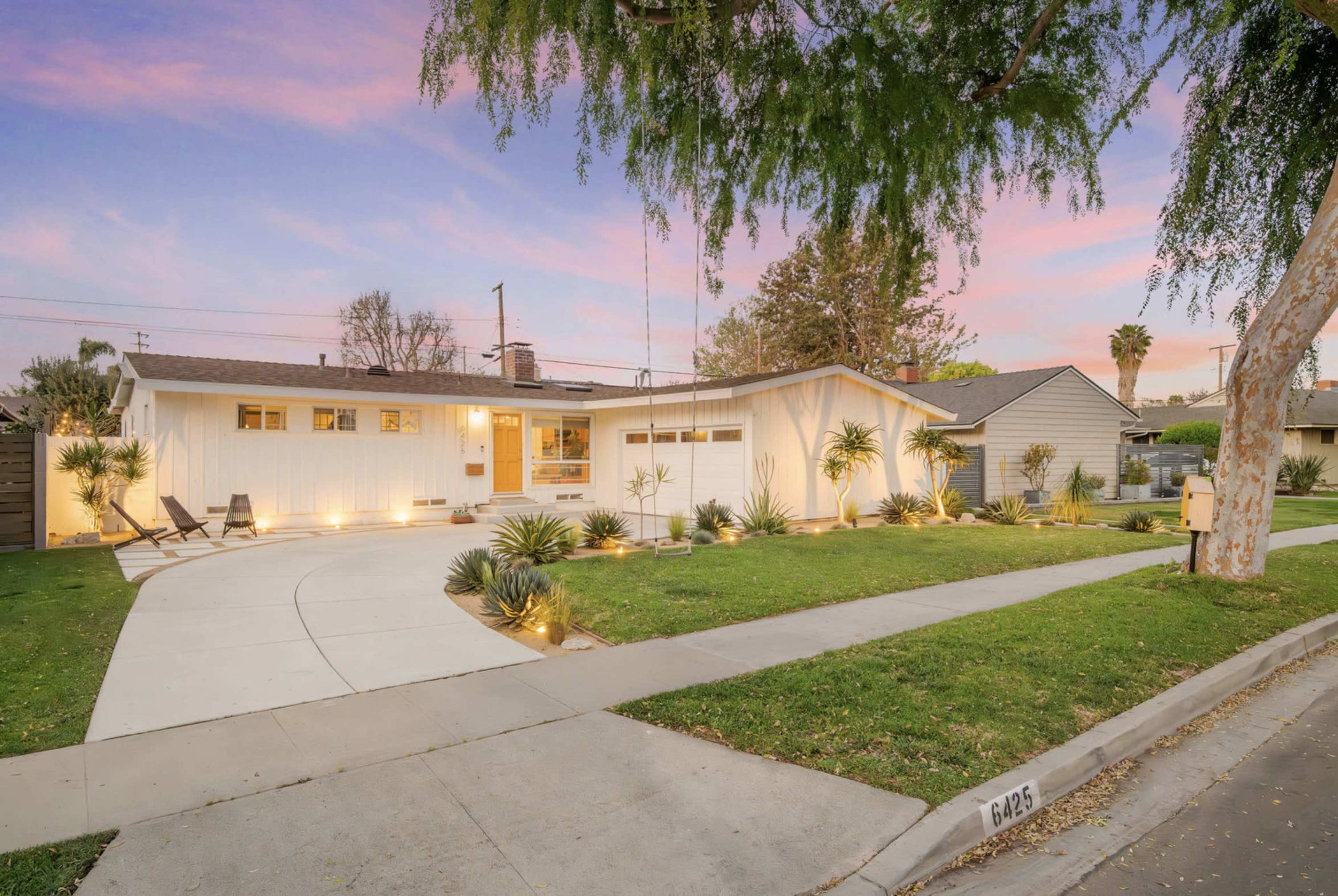 A single-story white home with a curved driveway, surrounded by landscaped greenery and illuminated at dusk.