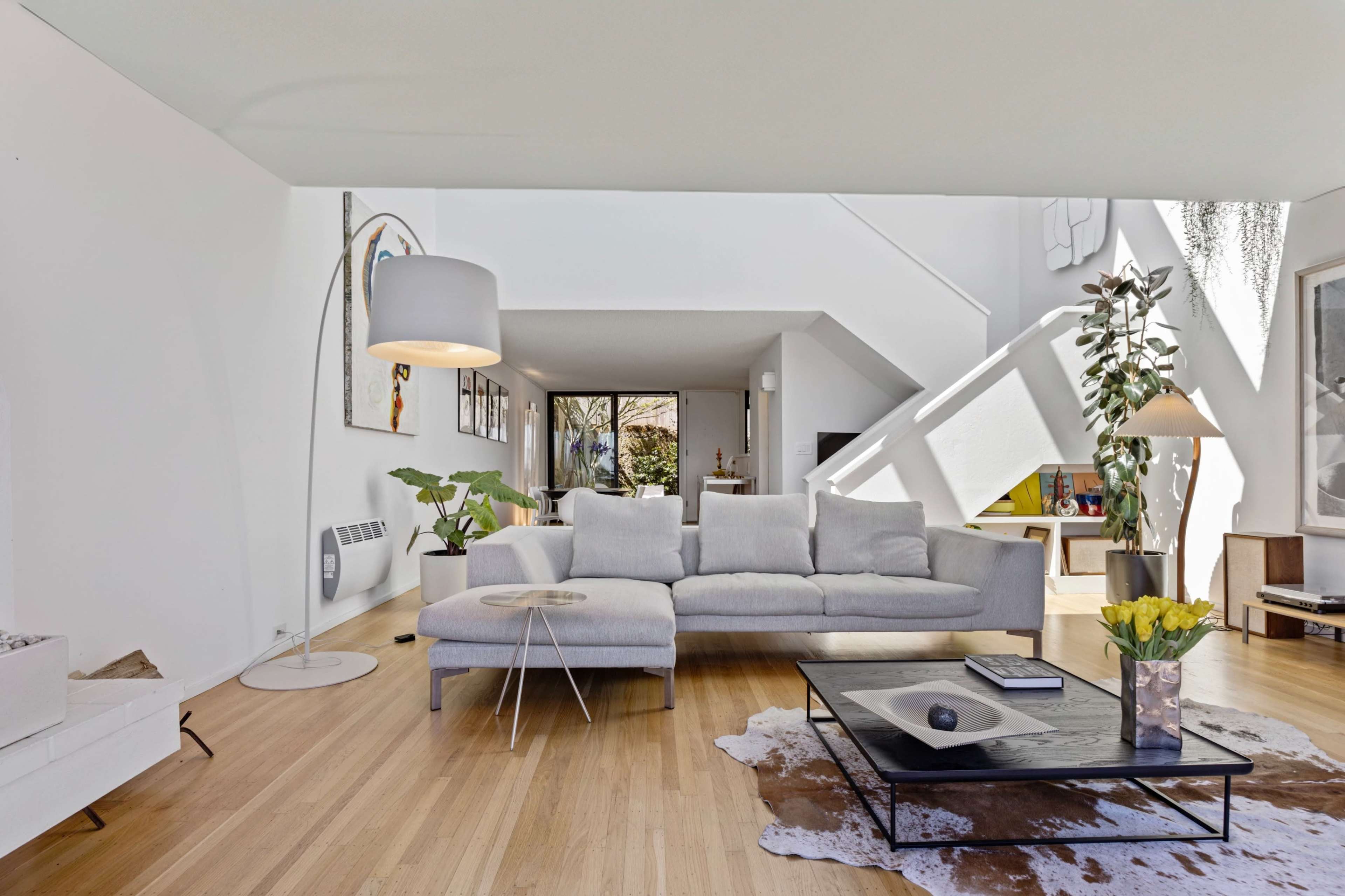 The image shows a modern living room featuring a light gray sectional sofa, a glass coffee table with a dark surface, and large windows allowing natural light to fill the space.
