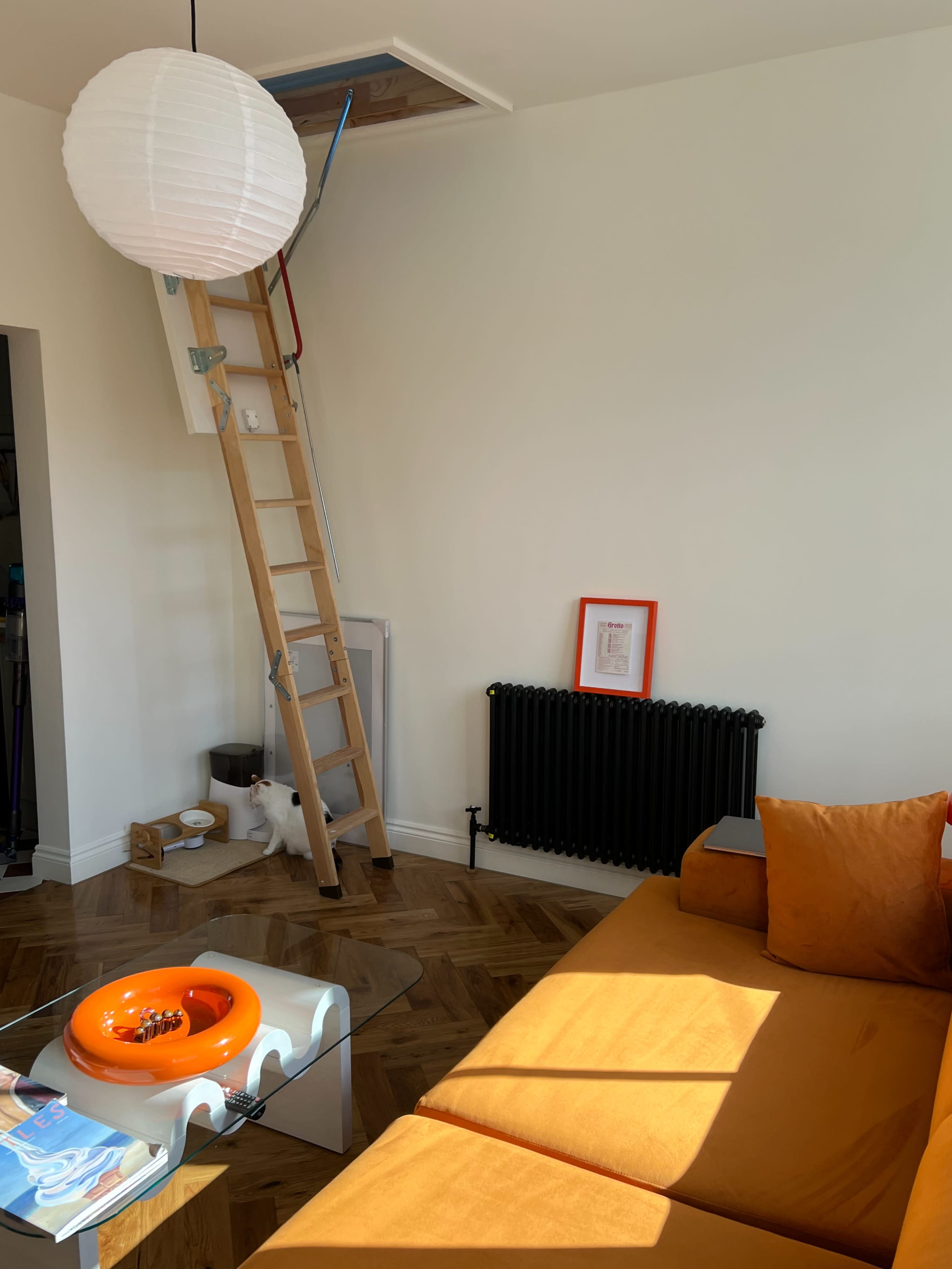 A wooden ladder leans against a wall beneath a ceiling hatch in a brightly lit room featuring an orange sofa and a radiator.