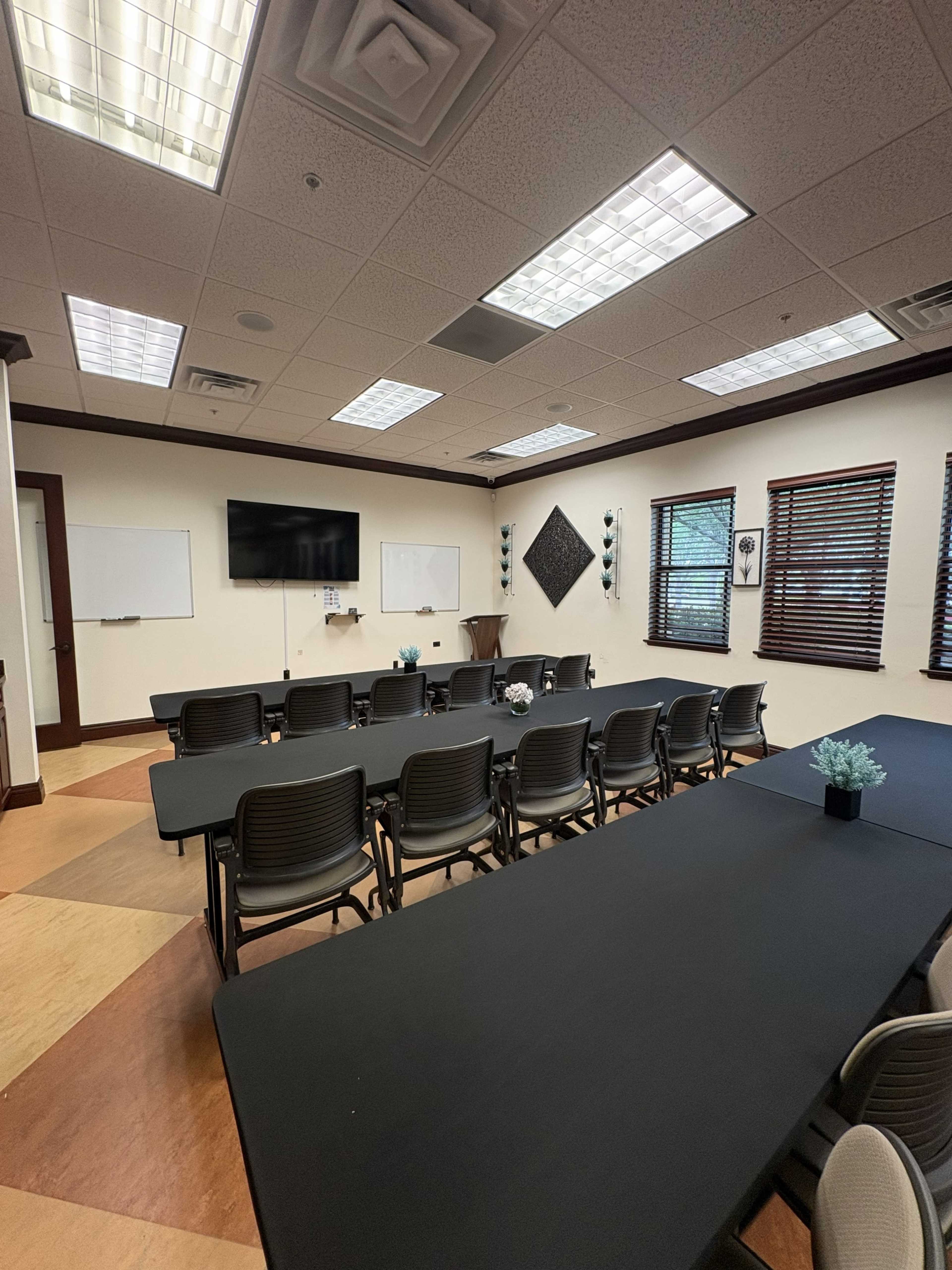 The image shows a conference room with multiple tables arranged in a U-shape, a large screen on the wall, and windows with blinds.