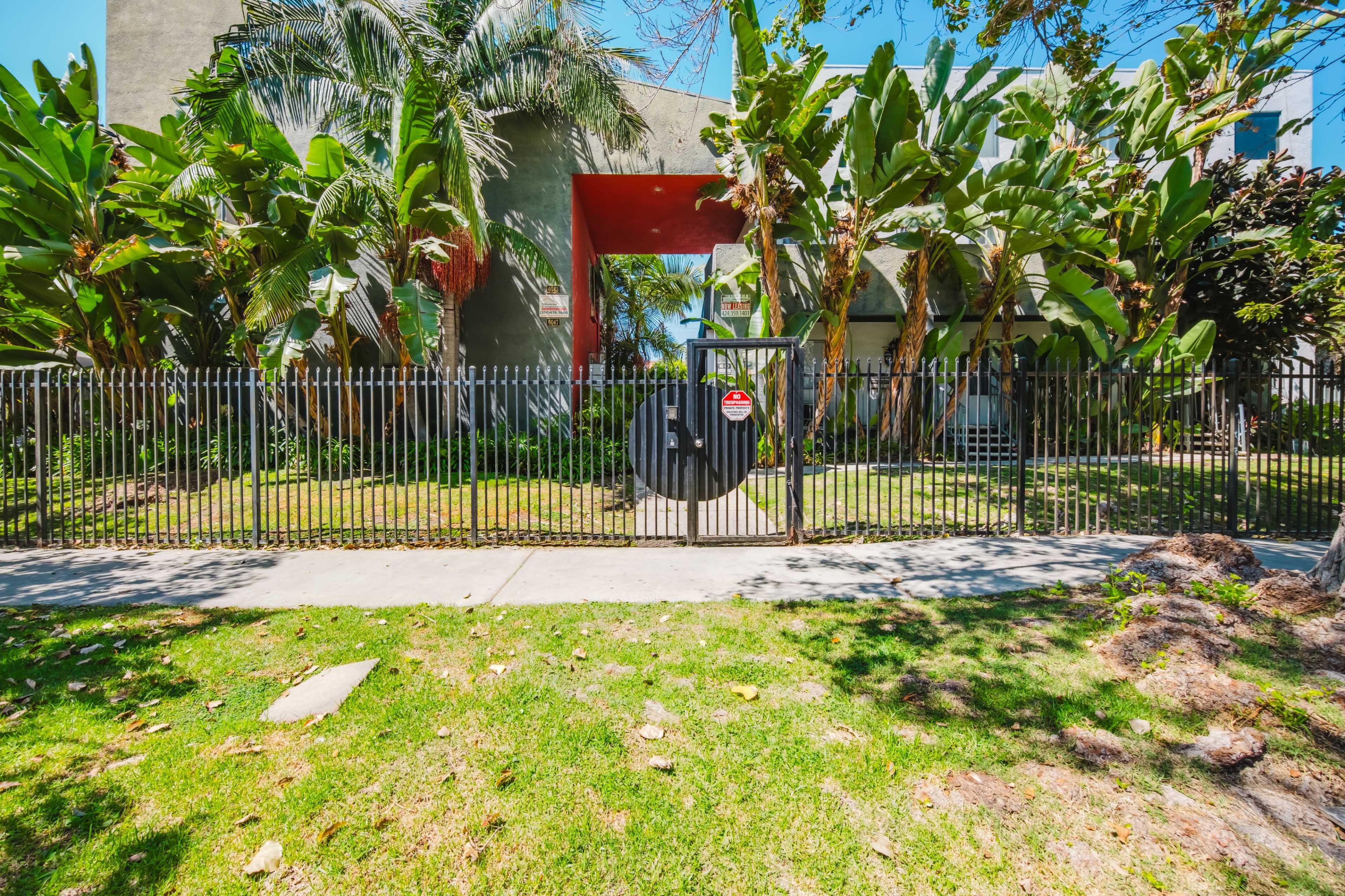 The image shows a gated entrance surrounded by lush greenery and palm trees, leading to a building in the background.