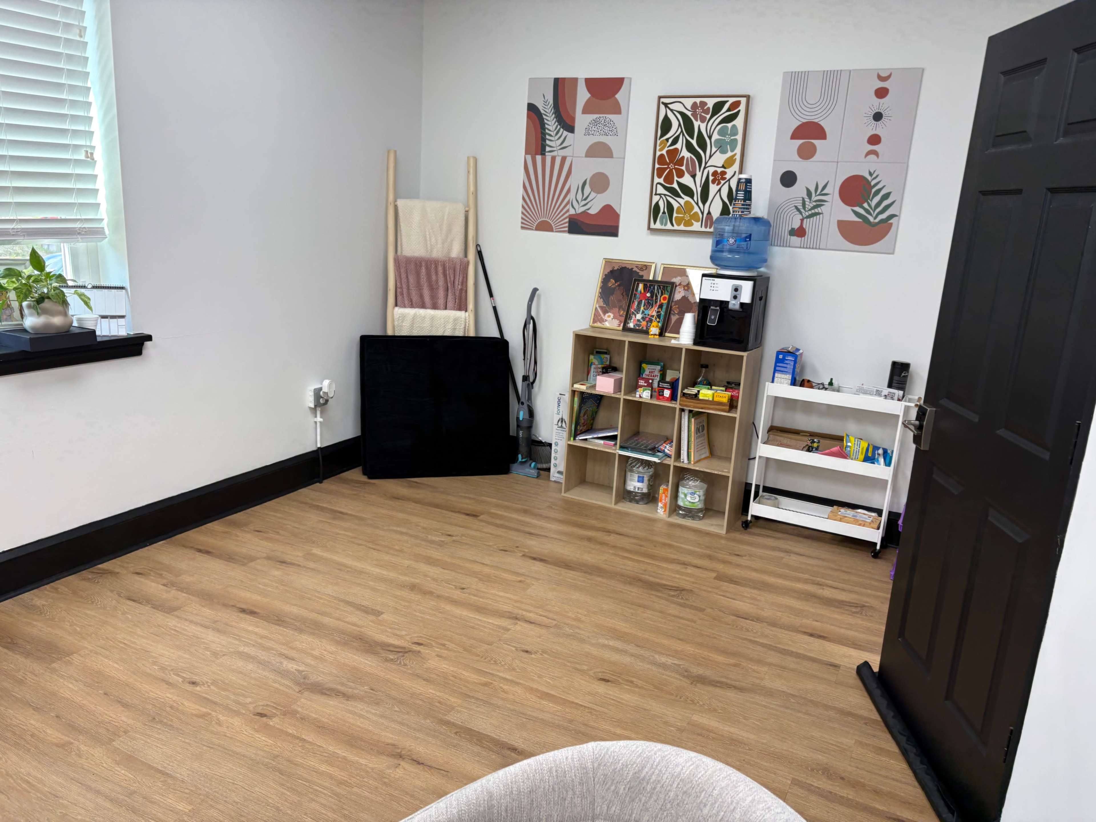 The image shows a well-lit room with light wood flooring, a shelf filled with various items, a water dispenser, a black chair, and two decorative wall art pieces.