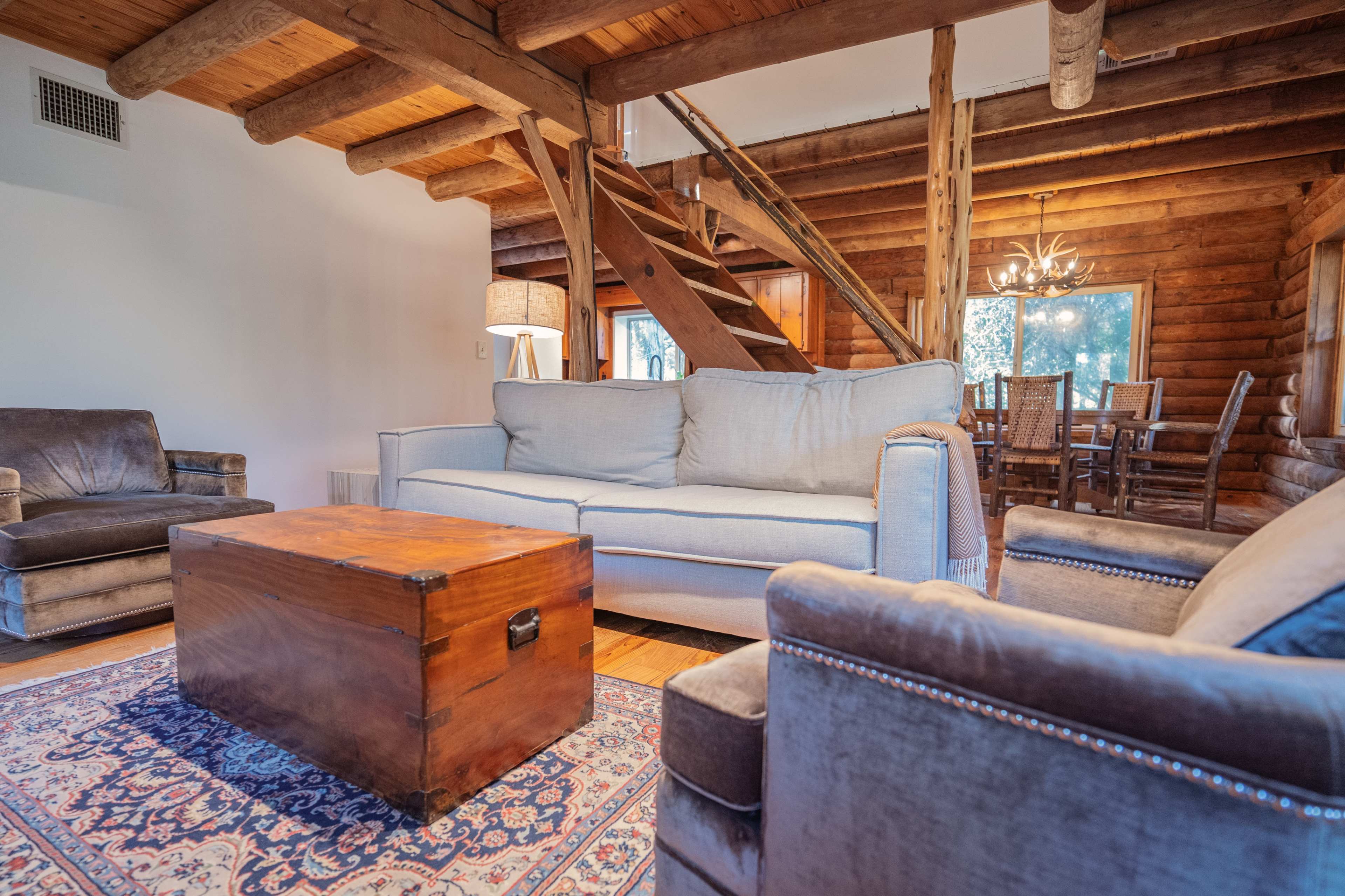 A cozy living room features a wooden beam ceiling, a light-colored sofa, a brown upholstered armchair, and a wooden coffee table on a patterned rug, with a staircase leading to a dining area in the background.