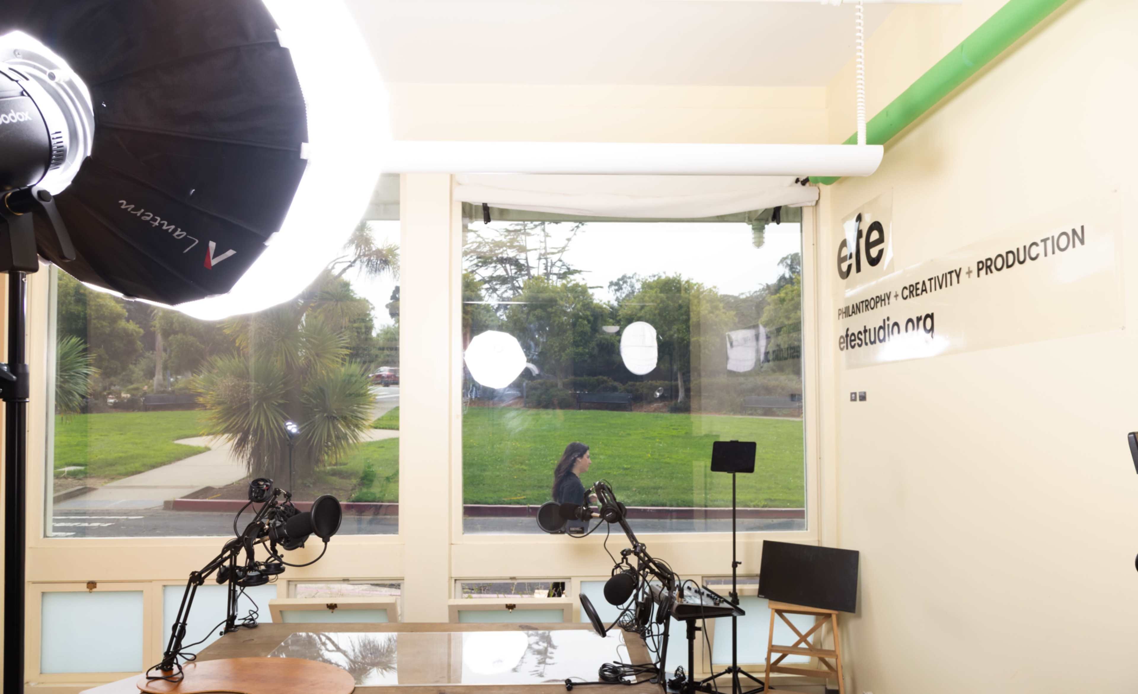 The image shows a brightly lit studio with large windows, featuring two microphones on stands, a camera lighting setup, and a table with a chair.