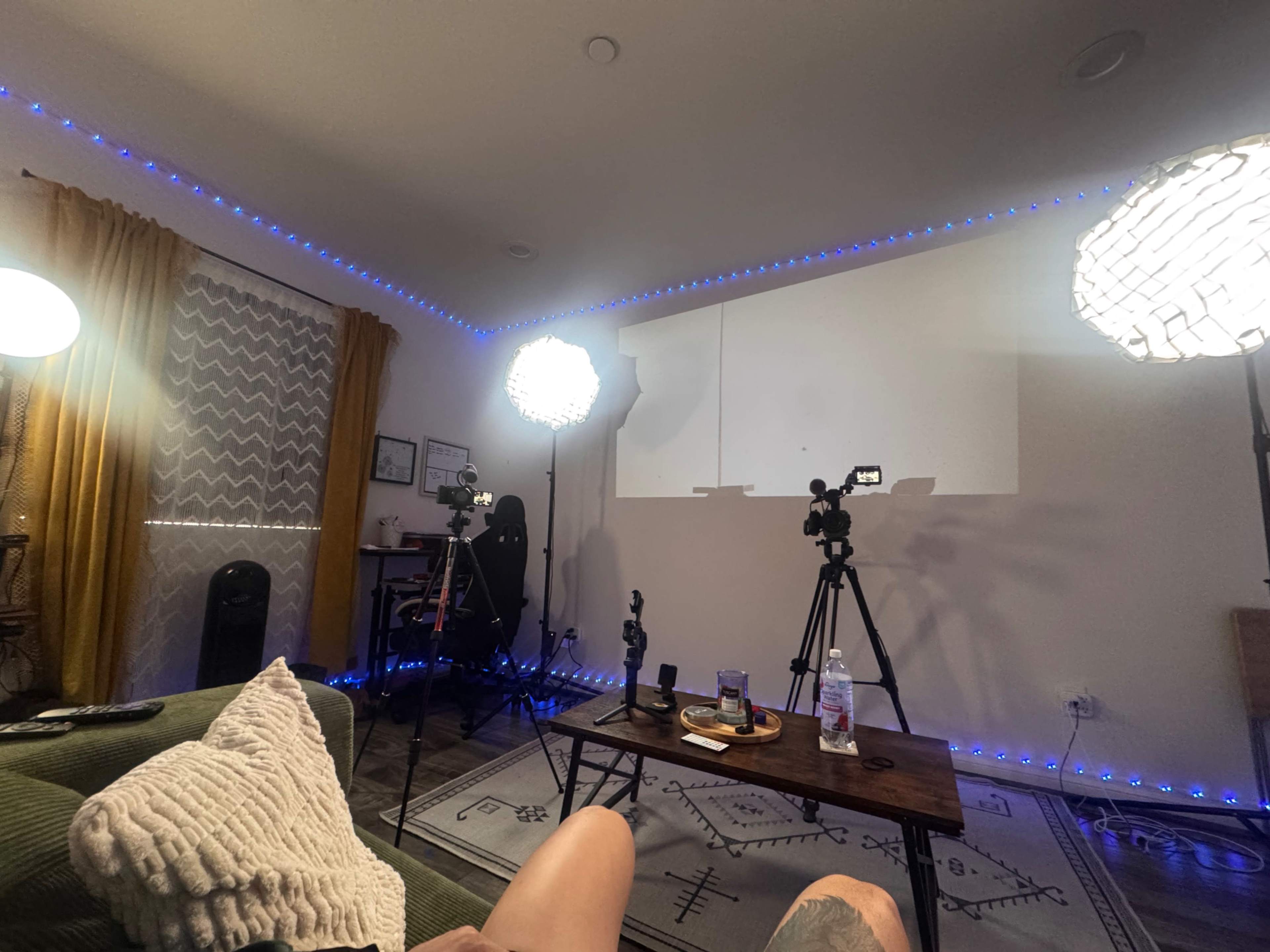 The image shows a well-lit room with two studio lights and two cameras set up facing a white projection screen, along with a couch and a low coffee table.