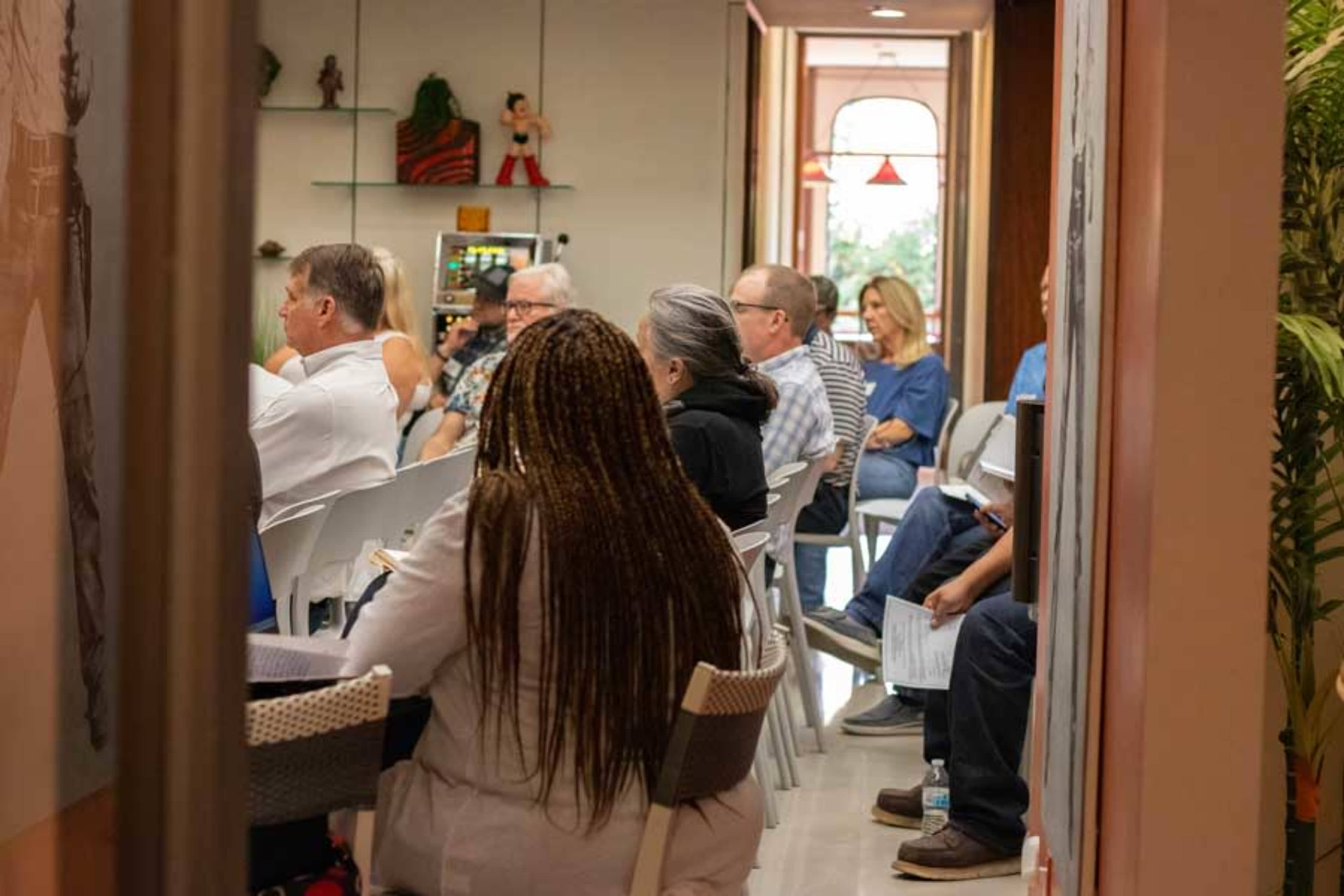 The image shows a group of people seated in a meeting room, facing forward, with various individuals engaged in discussion or listening attentively.