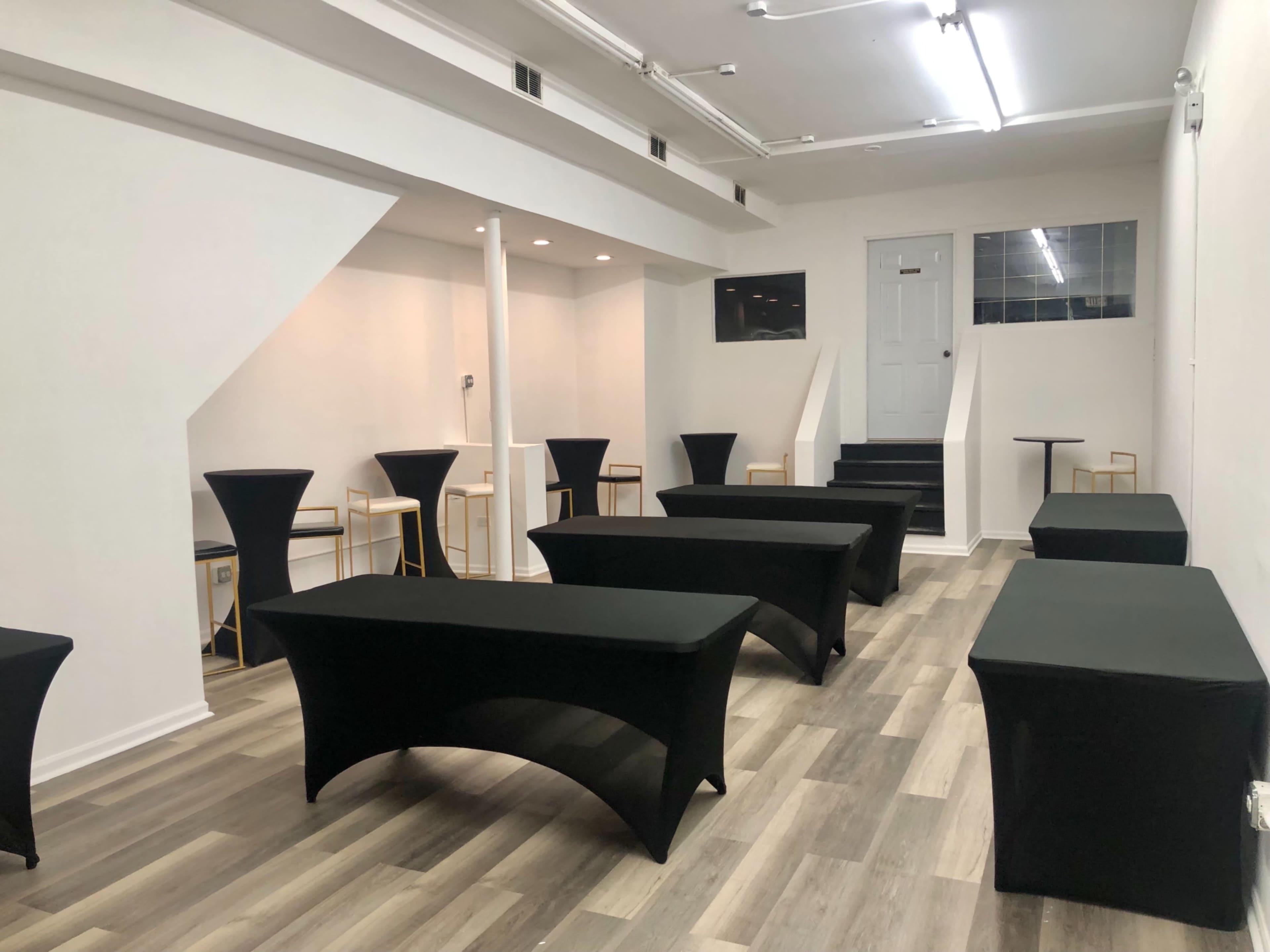 The image shows a spacious, empty room set up with several black-covered tables and a staircase at the far end.