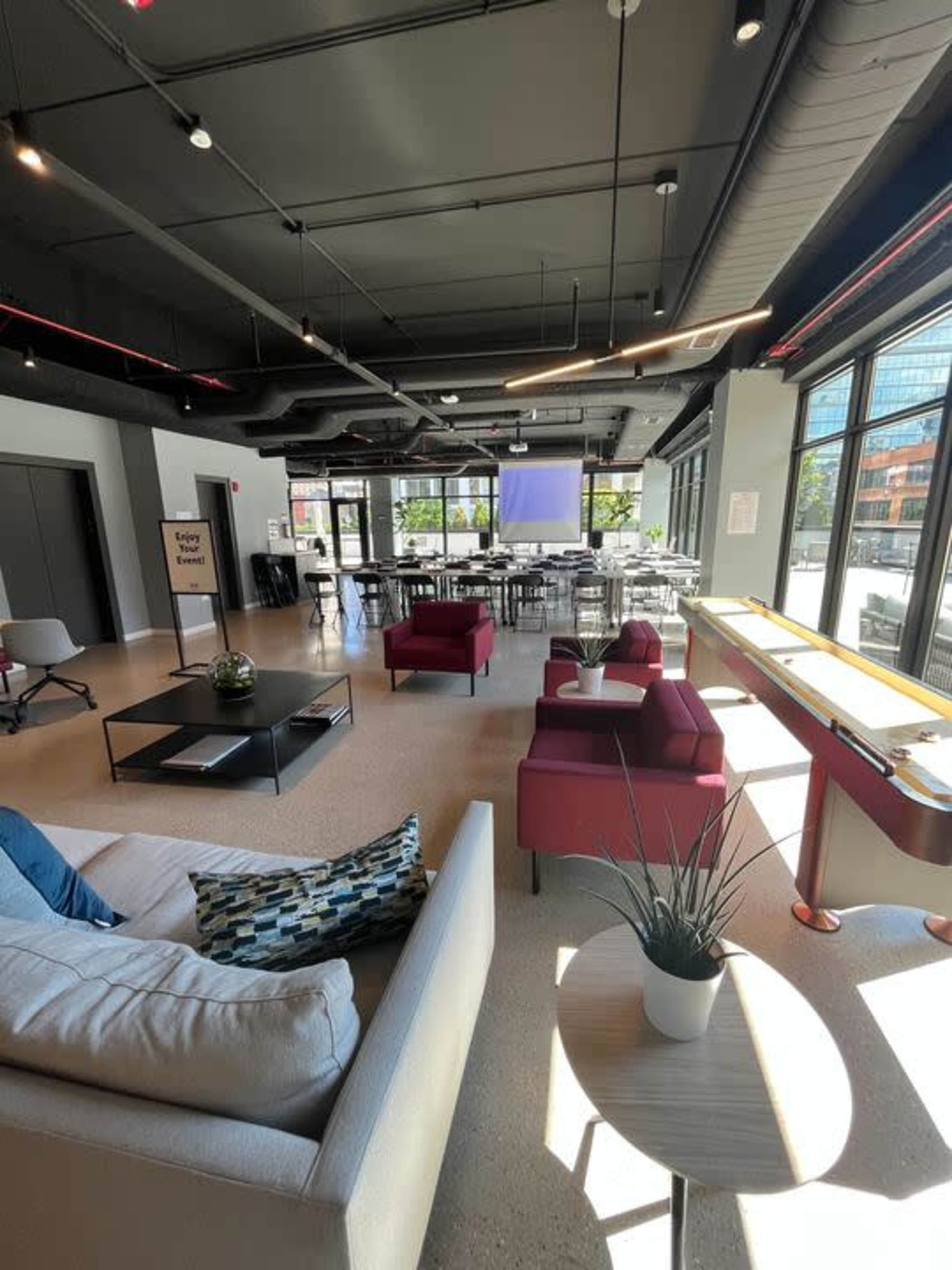 The image shows a spacious, modern lounge area with seating, tables, and large windows allowing natural light in.