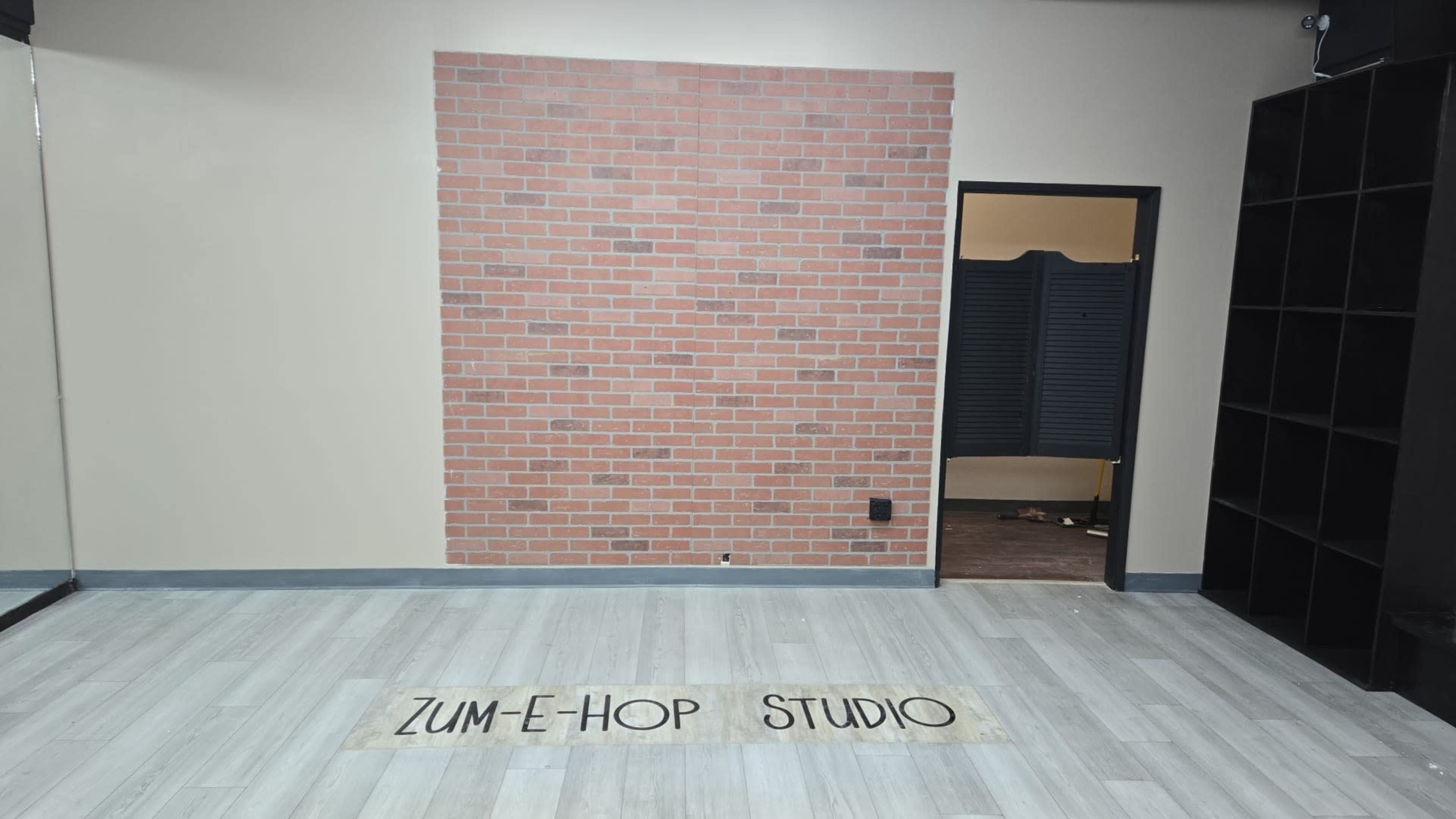 The image shows a dance studio with a brick-patterned accent wall, a mirrored wall, and a door leading to another room.