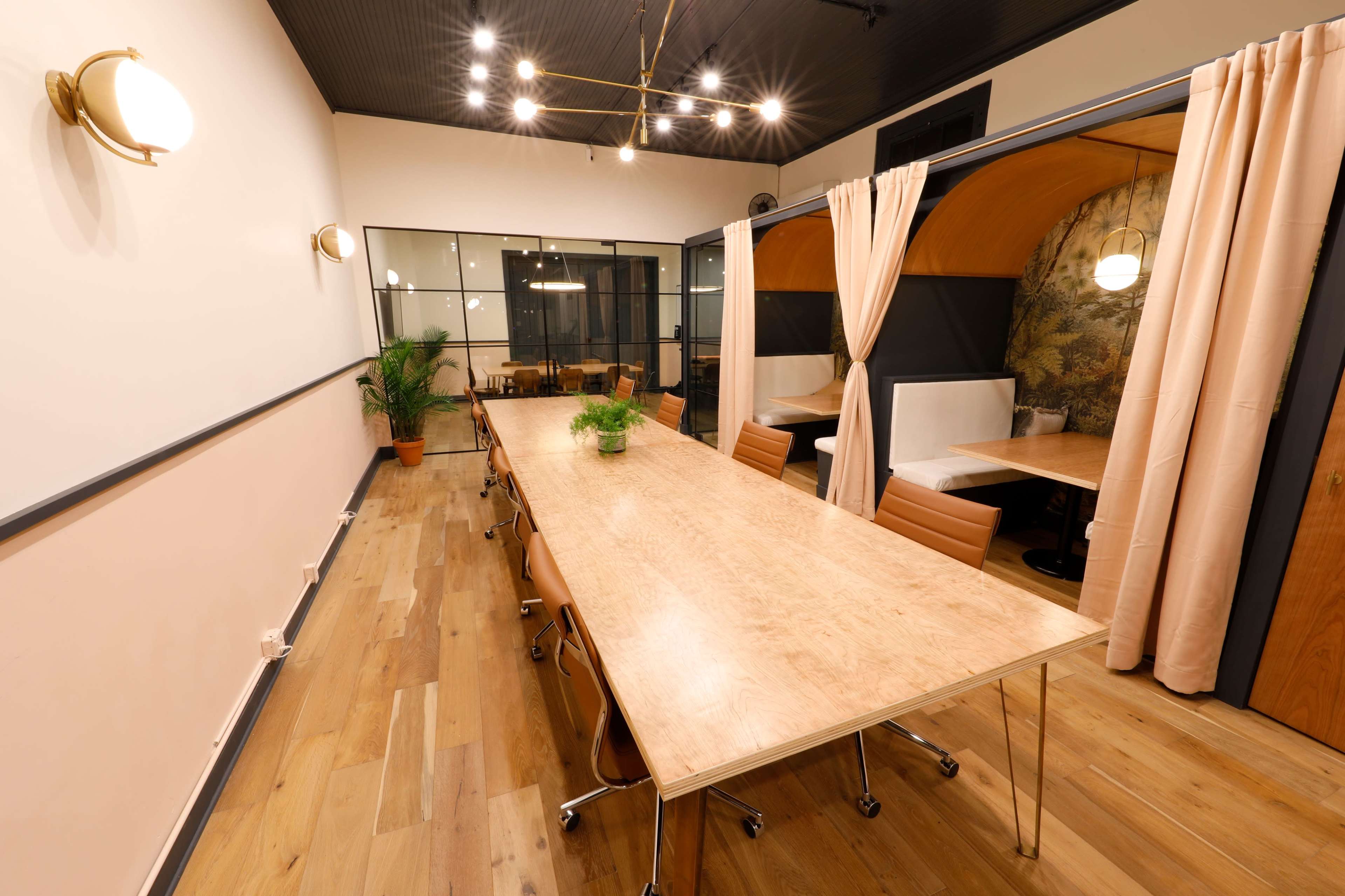 A large wooden table with rolling chairs is positioned in a well-lit meeting room featuring soft curtains and booths in the background.