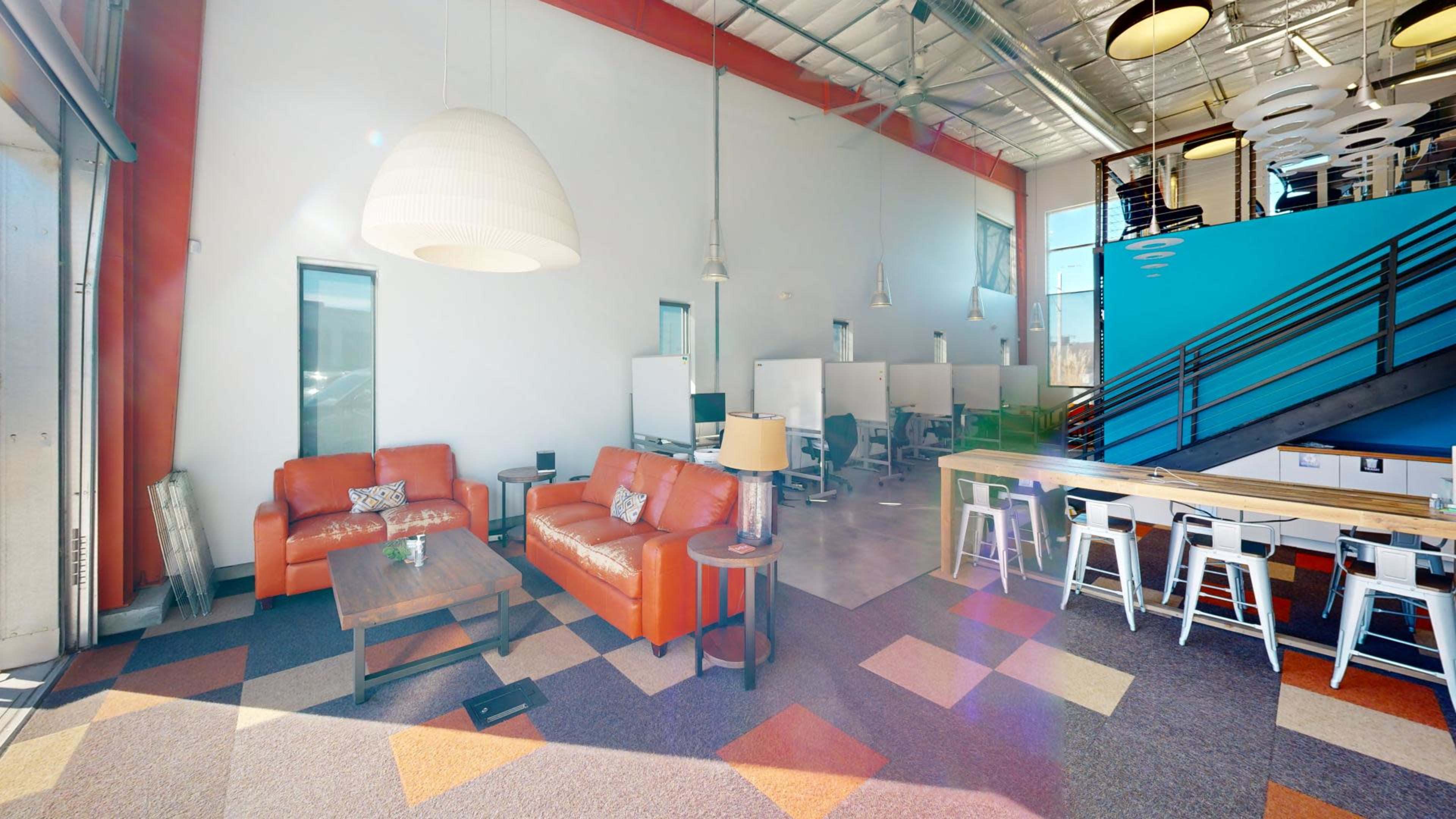 The image shows a modern office space featuring a lounge area with orange sofas, a coffee table, and a workspace with desks and chairs.